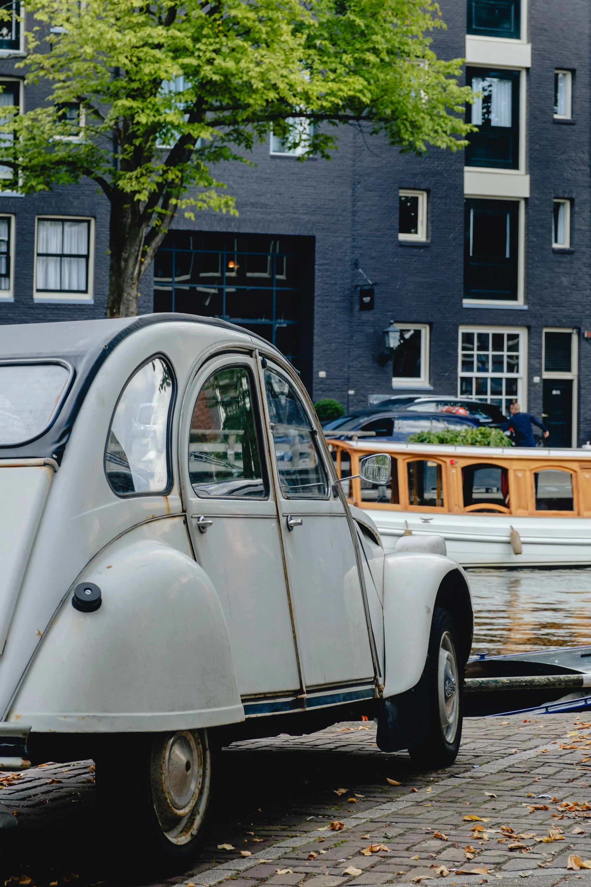 An old white vintage car parked on a cobblestone street near a canal, with a modern black apartment building and a tree in the background.