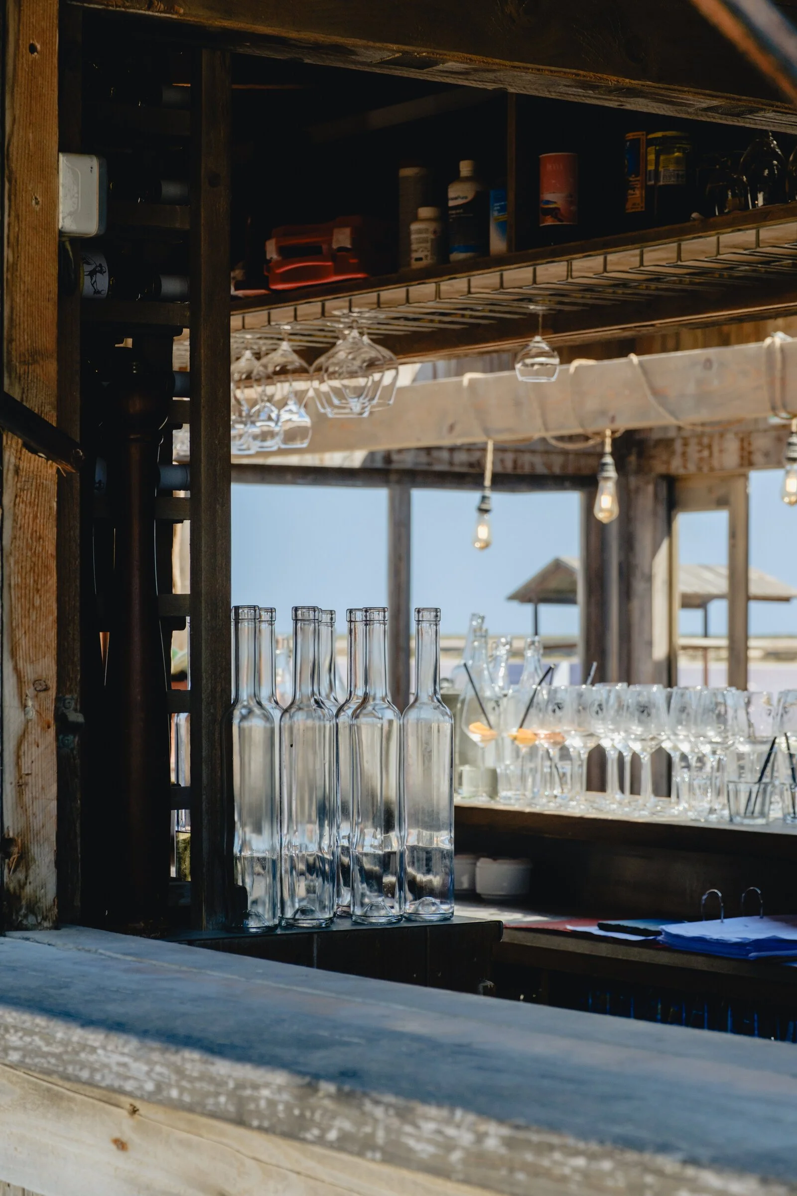 Glass bottles and stemware line a rustic wooden beach bar, string lights strung overhead