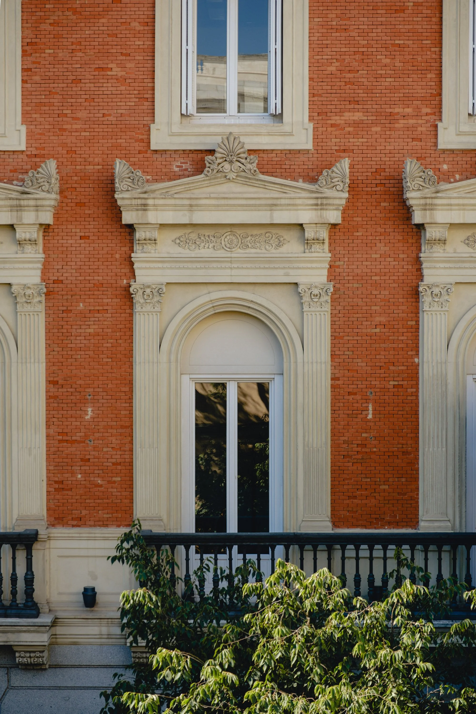 Close-up of a historic building facade with red brick walls, decorative white stone window frames, and ornate architectural details above the windows. Green foliage is visible at the bottom.
