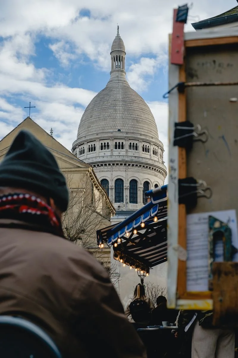 The white dome of Sacré-Cœur rises behind a lit market stall and passersby in Montmartre