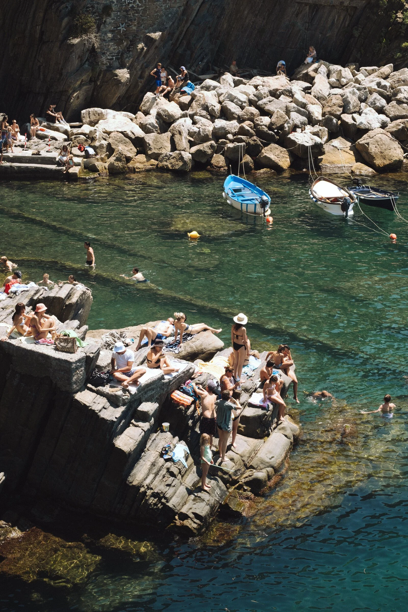 Sunbathers spread across flat cliffside rocks above turquoise water, swimmers and small boats nearby