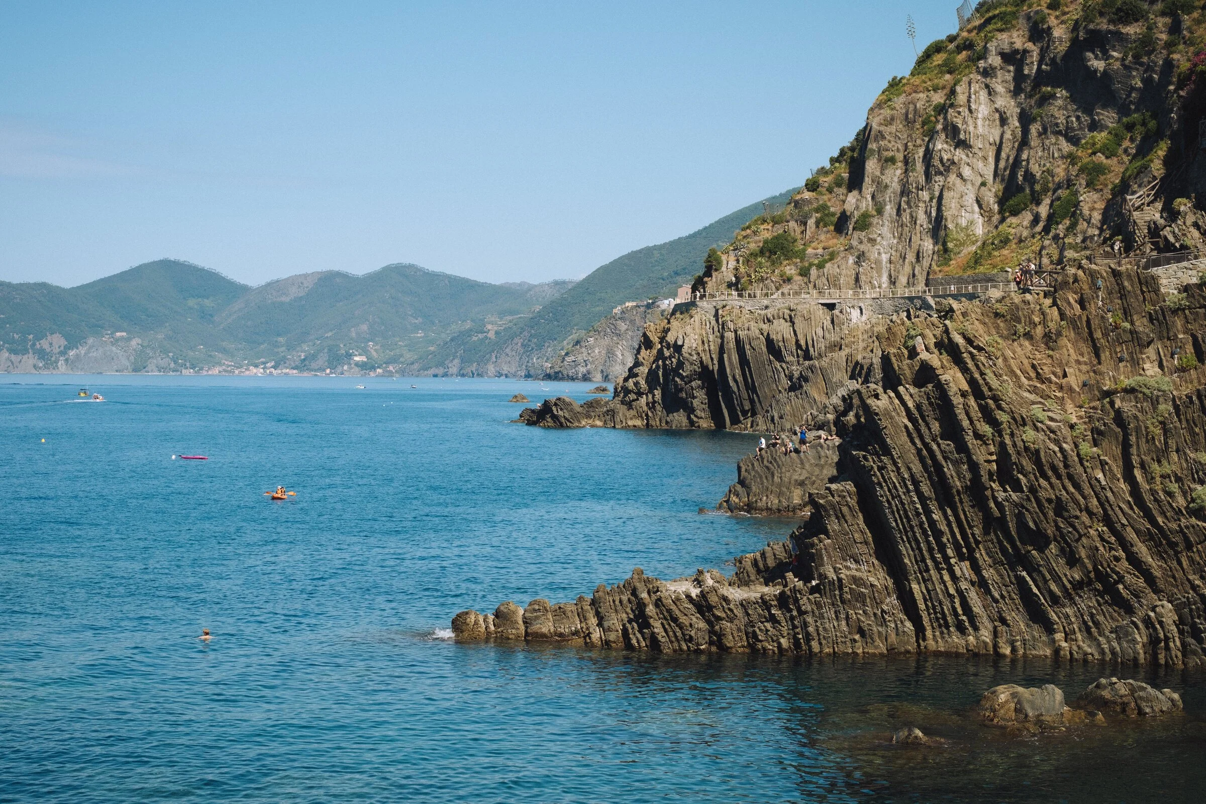 A terraced cliff path above a turquoise cove in Riomaggiore, swimmers below and boats moored offshore