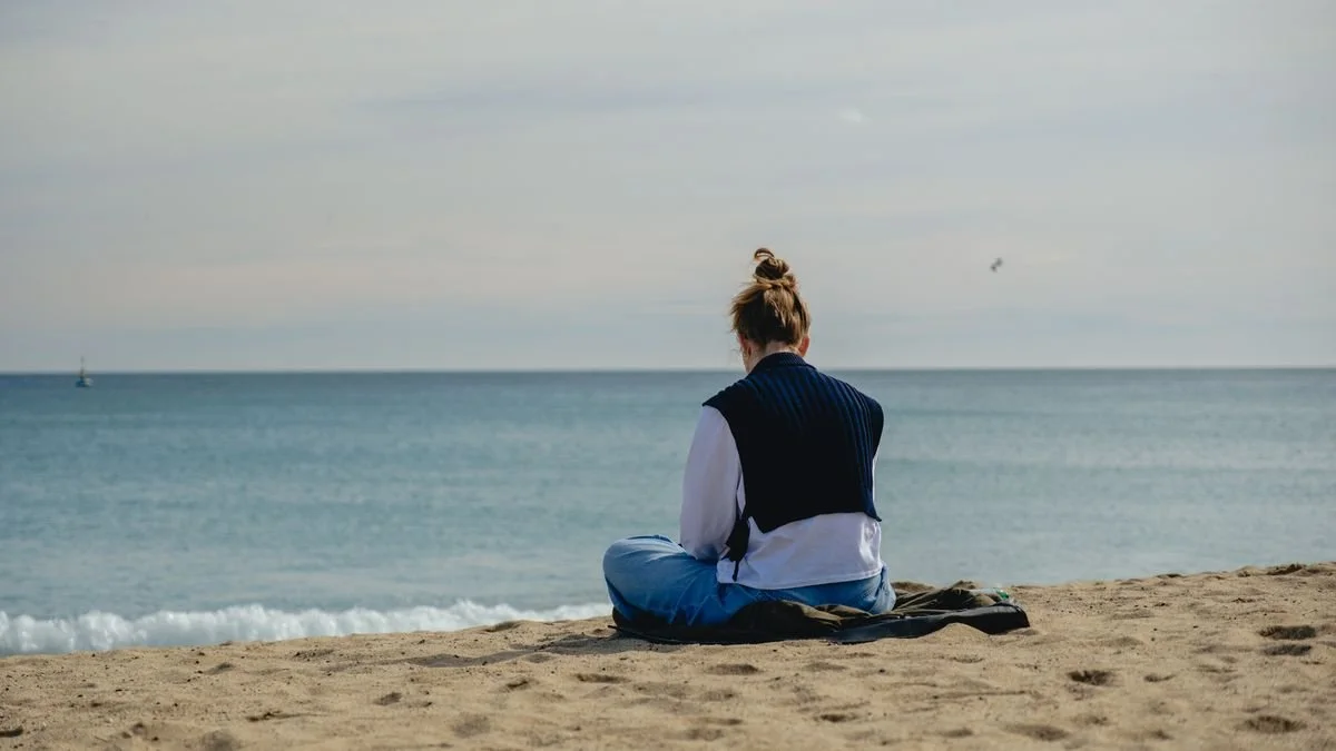 A woman sits cross-legged on the sand, facing the sea, her back to the camera
