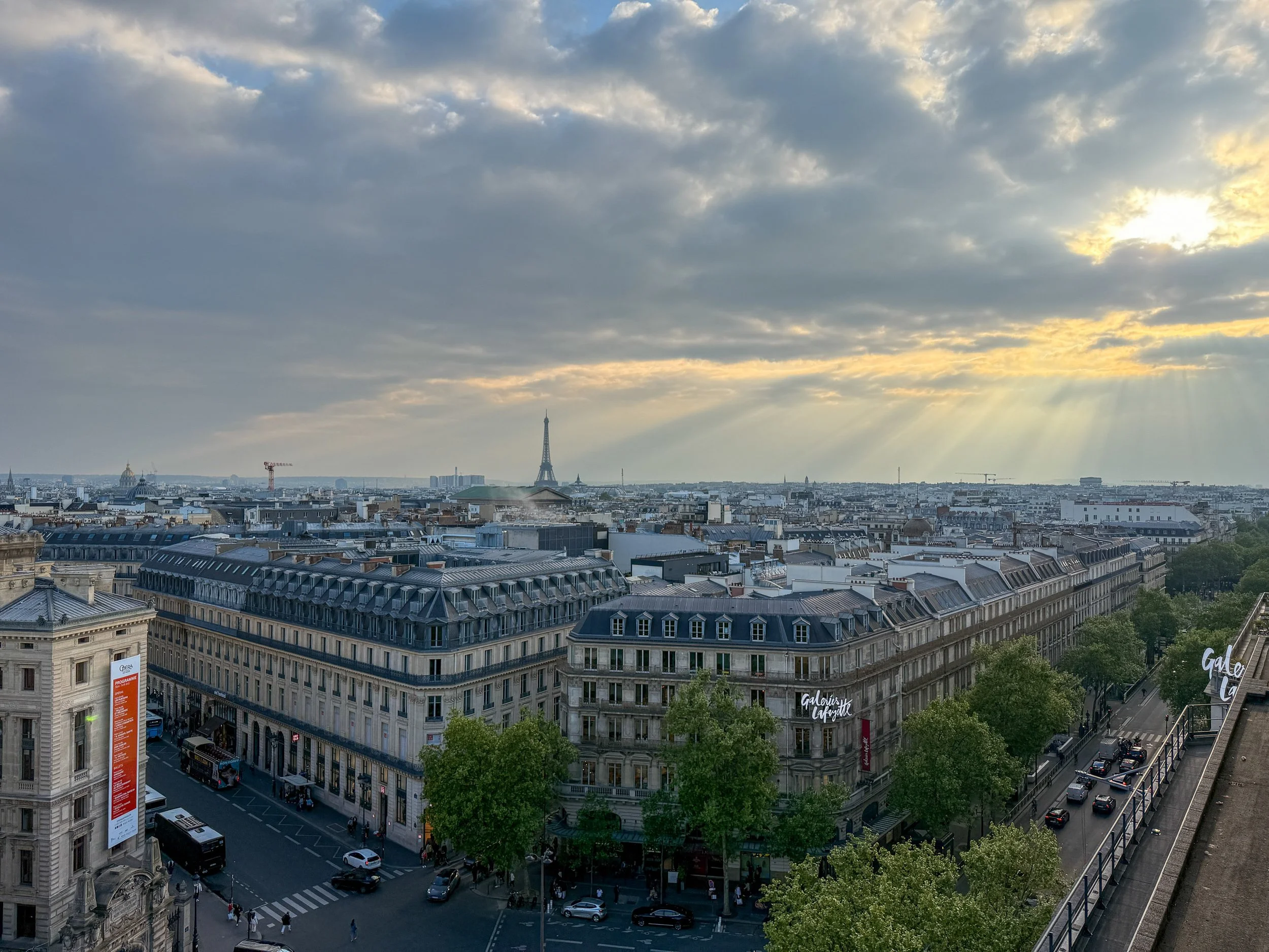 Cityscape view of Paris with the Eiffel Tower in the distance, buildings, trees, and a cloudy sky with rays of sunlight breaking through.
