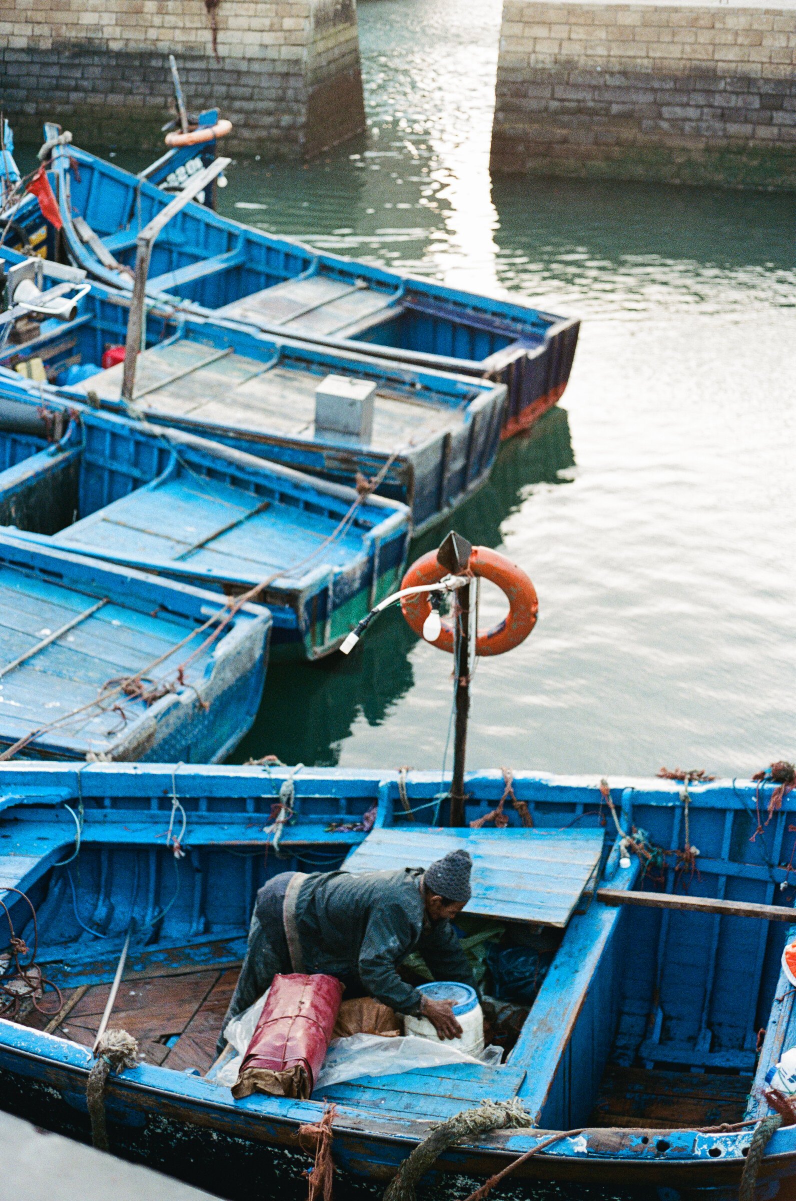 A fisherman crouches sorting his gear among a tangle of blue wooden boats