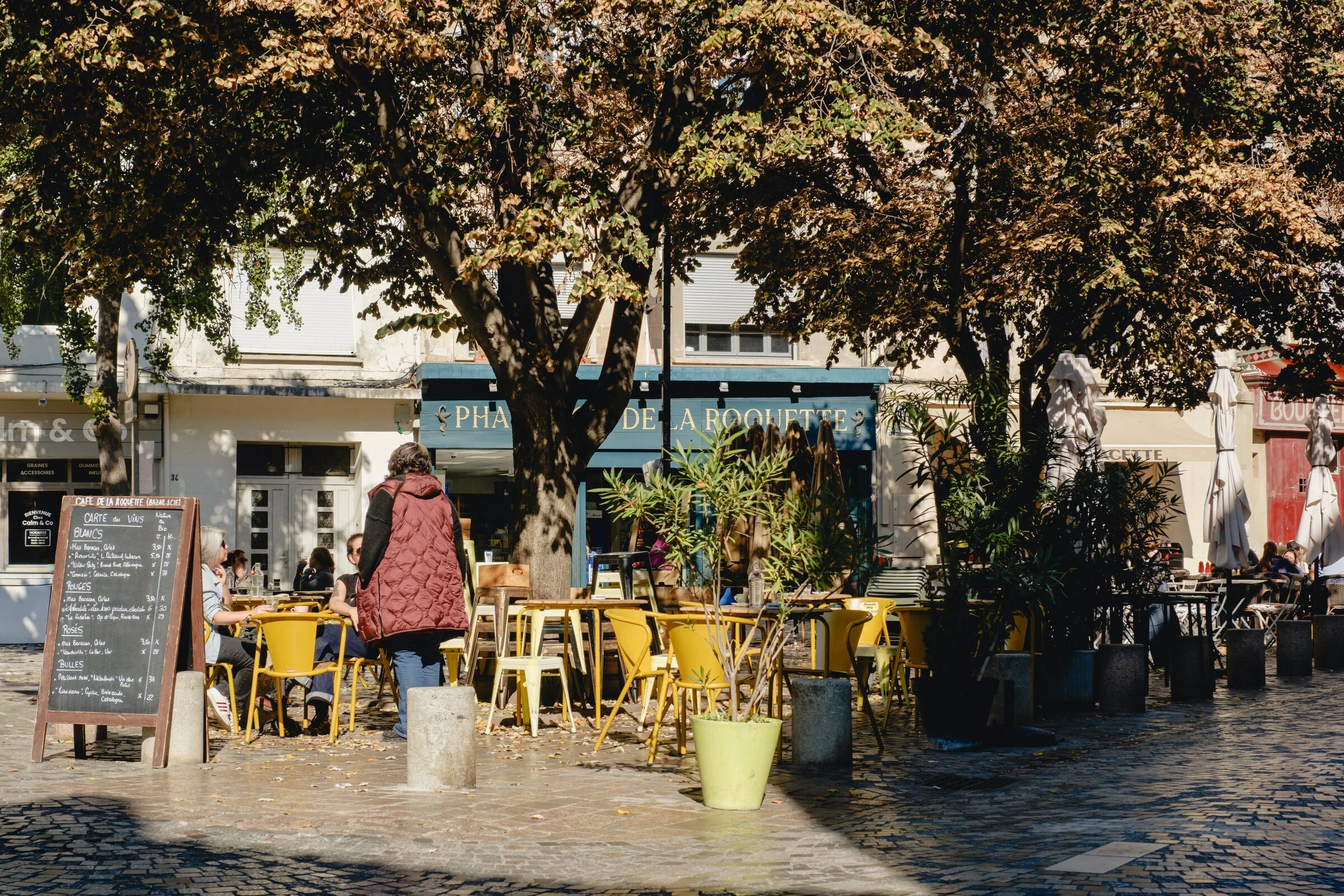 Yellow bistro chairs cluster under shade trees outside a café on a town square