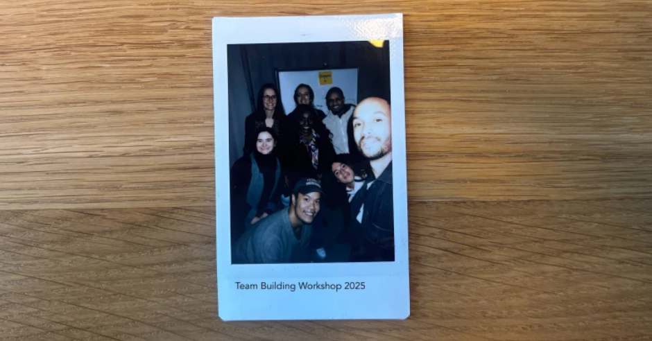 Fotografie einer Gruppe von acht Menschen während eines Team Building Workshops 2025, aufgenommen im Innenraum auf einem Holztisch.
