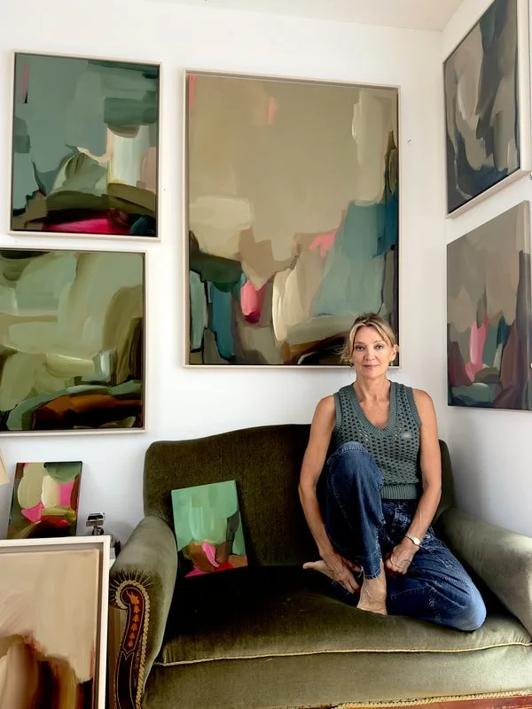 Carrie Jean Goldsmith on a vintage green sofa surrounded by her paintings on the walls and leaning on the floor.