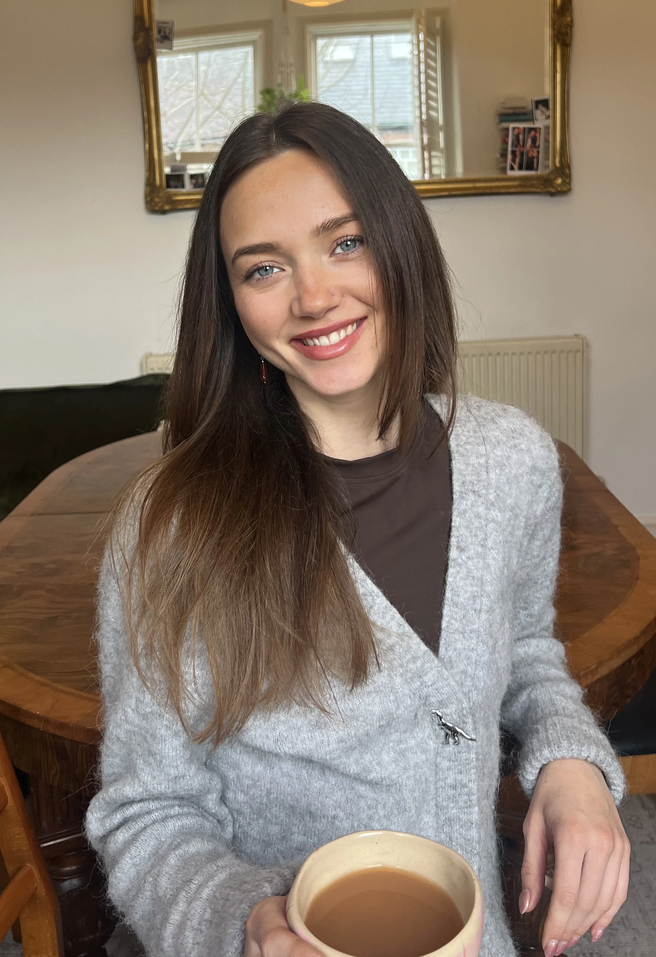 A young woman with long brown hair and blue eyes smiling and holding a mug of coffee or tea, standing in a cozy kitchen or dining area.