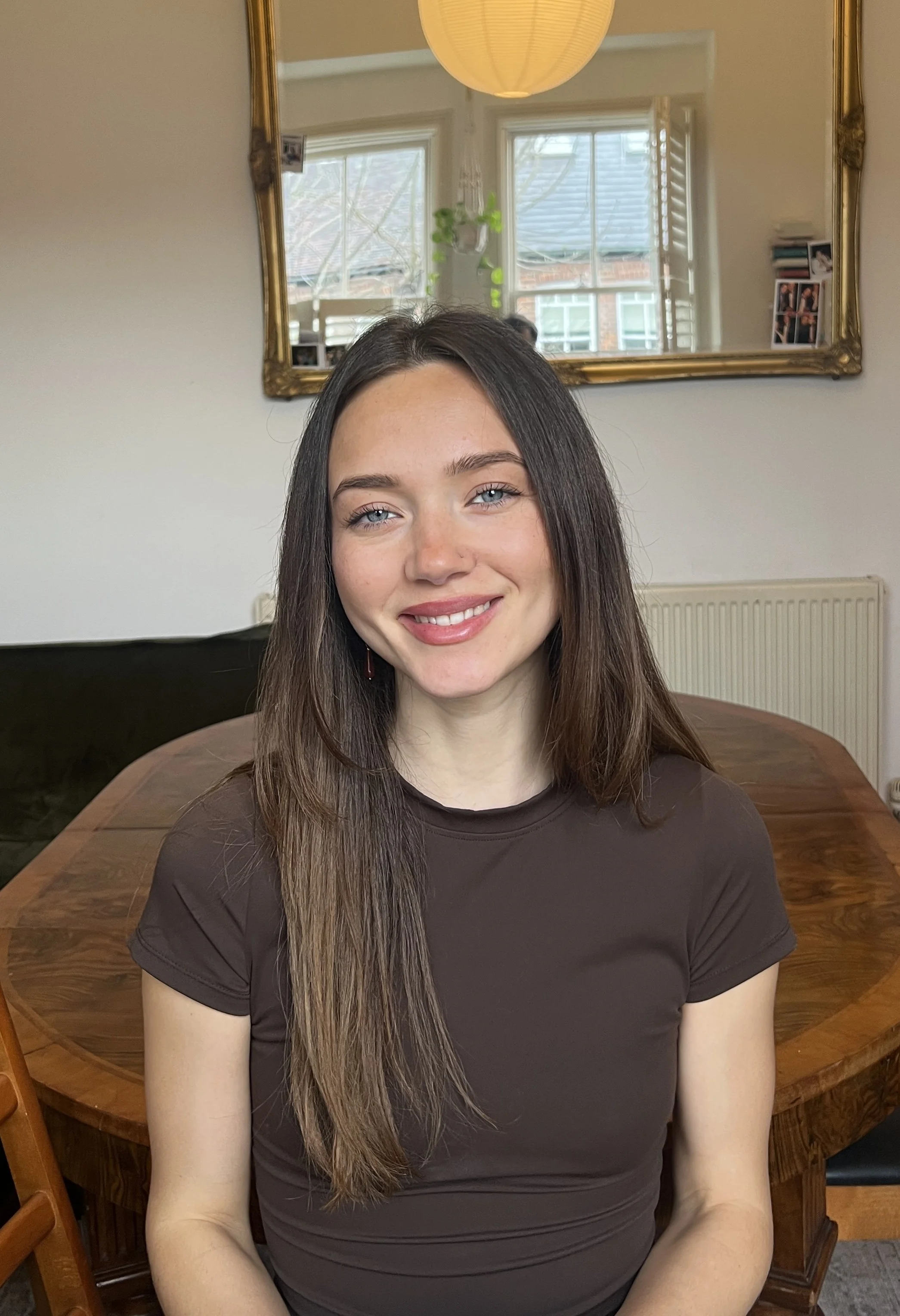 A woman with long brown hair, blue eyes, and wearing a dark brown shirt, smiling and sitting at a wooden table in a cozy home setting with a mirror, window, and pictures in the background.