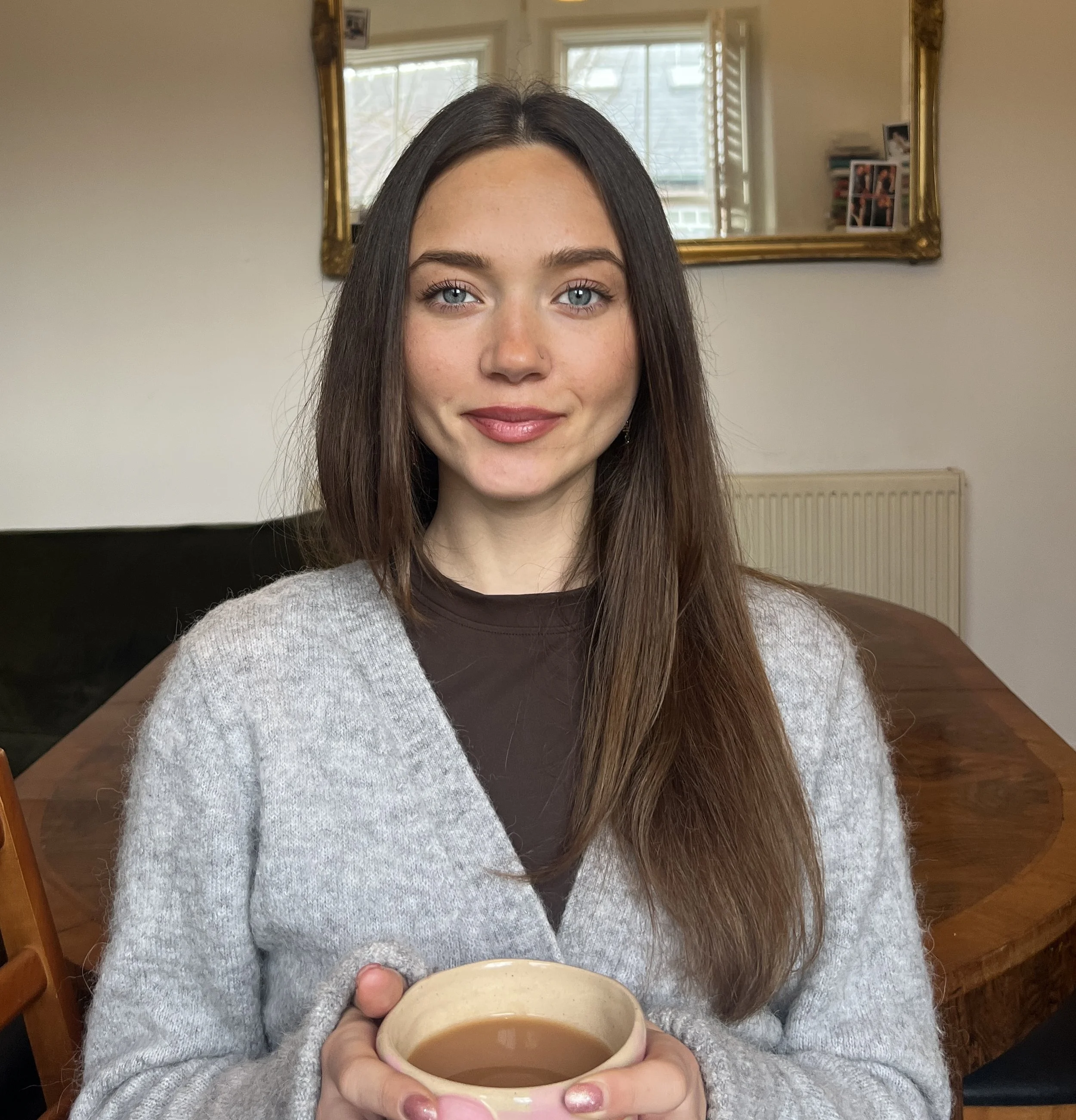 A young woman with long brown hair and blue eyes holding a cup of tea indoors, standing in front of a mirror and a wooden dining table.