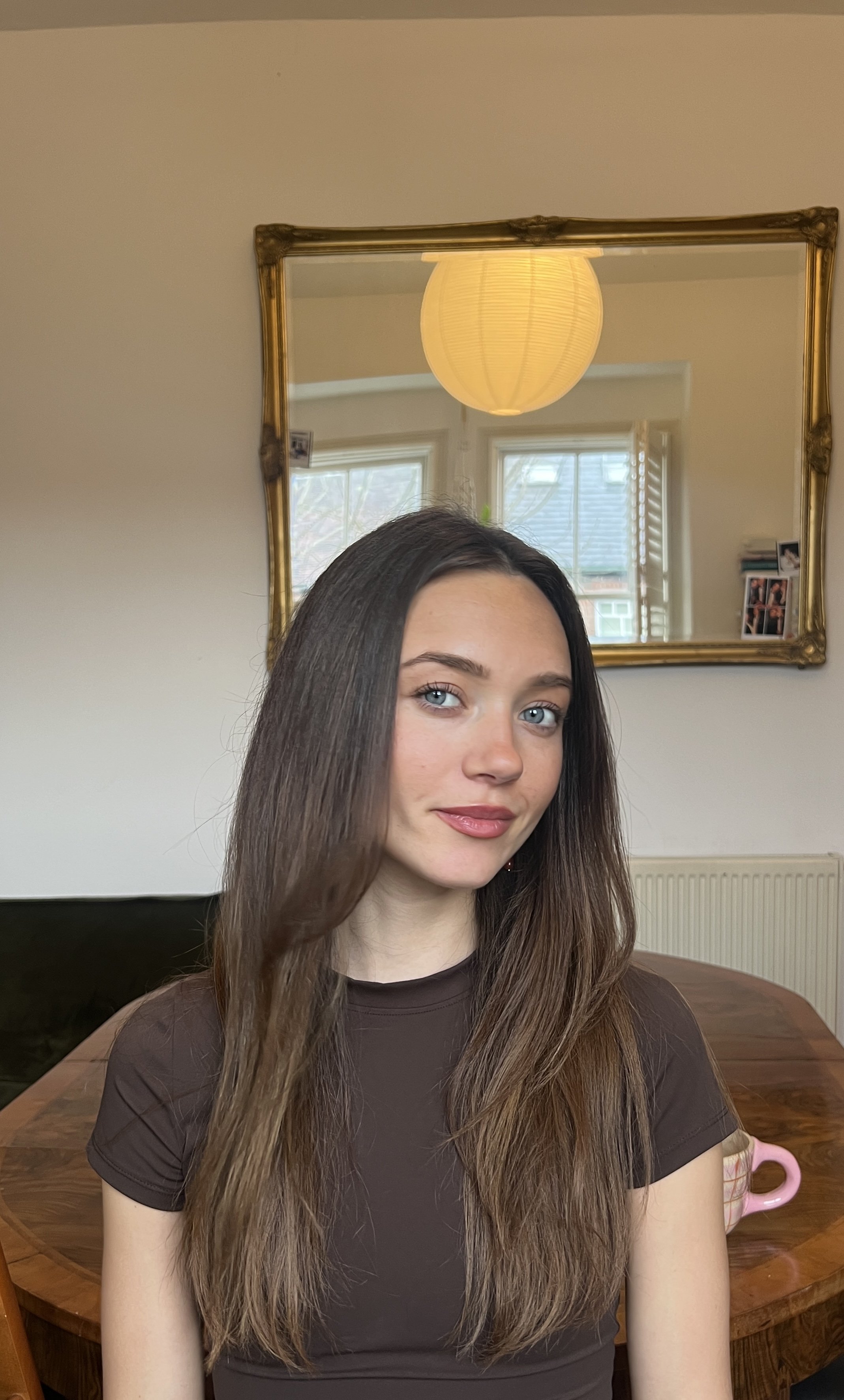 A young woman with long, wavy brown hair and blue eyes, wearing a dark brown shirt, sitting at a wooden dining table in a room with a large window, a mirror with a gold frame, a paper lantern ceiling light, and some photos on the windowsill.