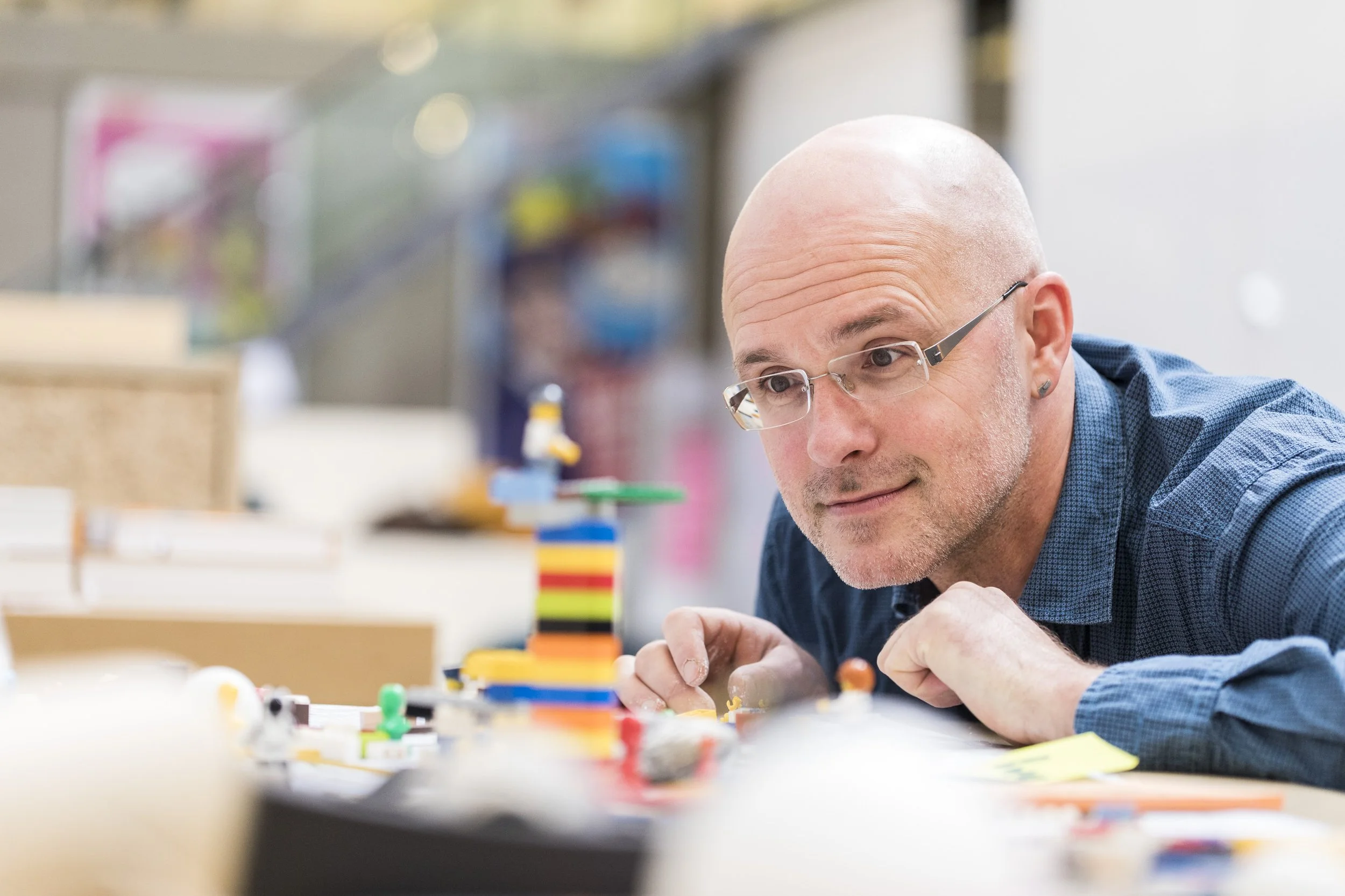 Ein Mann mit Brille und Kurzhaar, der an einem Tisch mit bunten Spielzeug oder Bauklötzen arbeitet, in einem gut beleuchteten Raum.