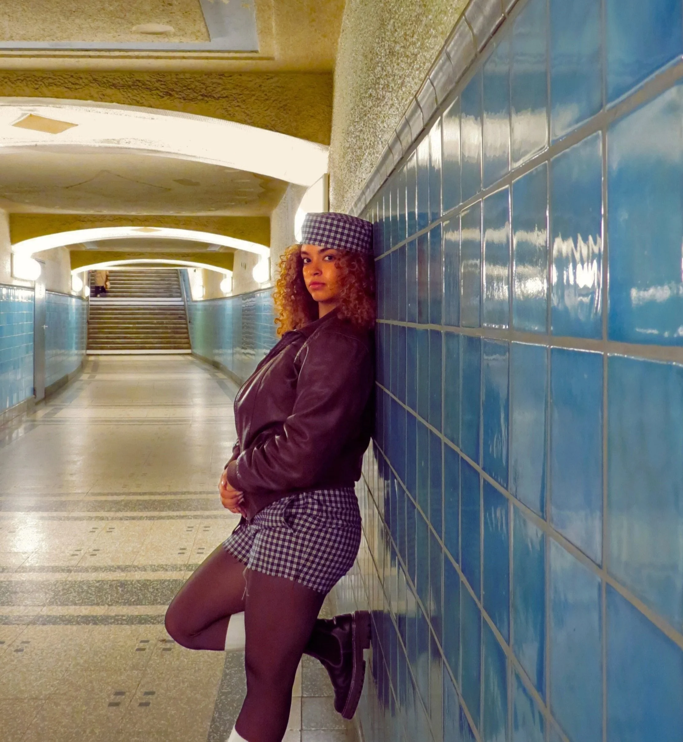 Sarah Edoghogho poses confidently in an underground tunnel with blue tiled walls and warm lighting, wearing a checkered outfit with matching cap, dark jacket, tights, and black boots.