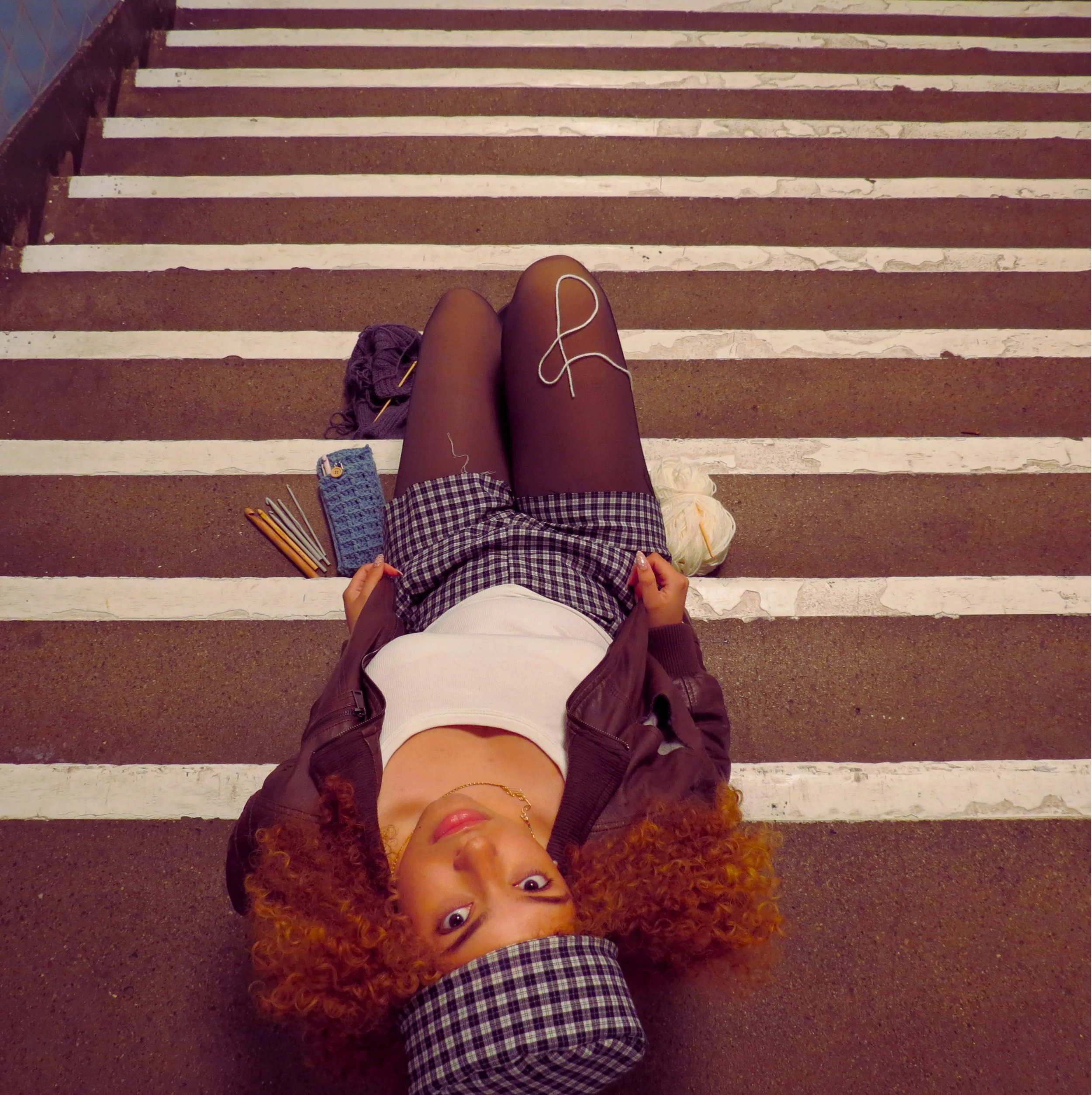 Sarah Edoghogho looking up  with crochet supplies around her wearing a checkered outfit with matching cap, dark jacket, tights, and black boots.