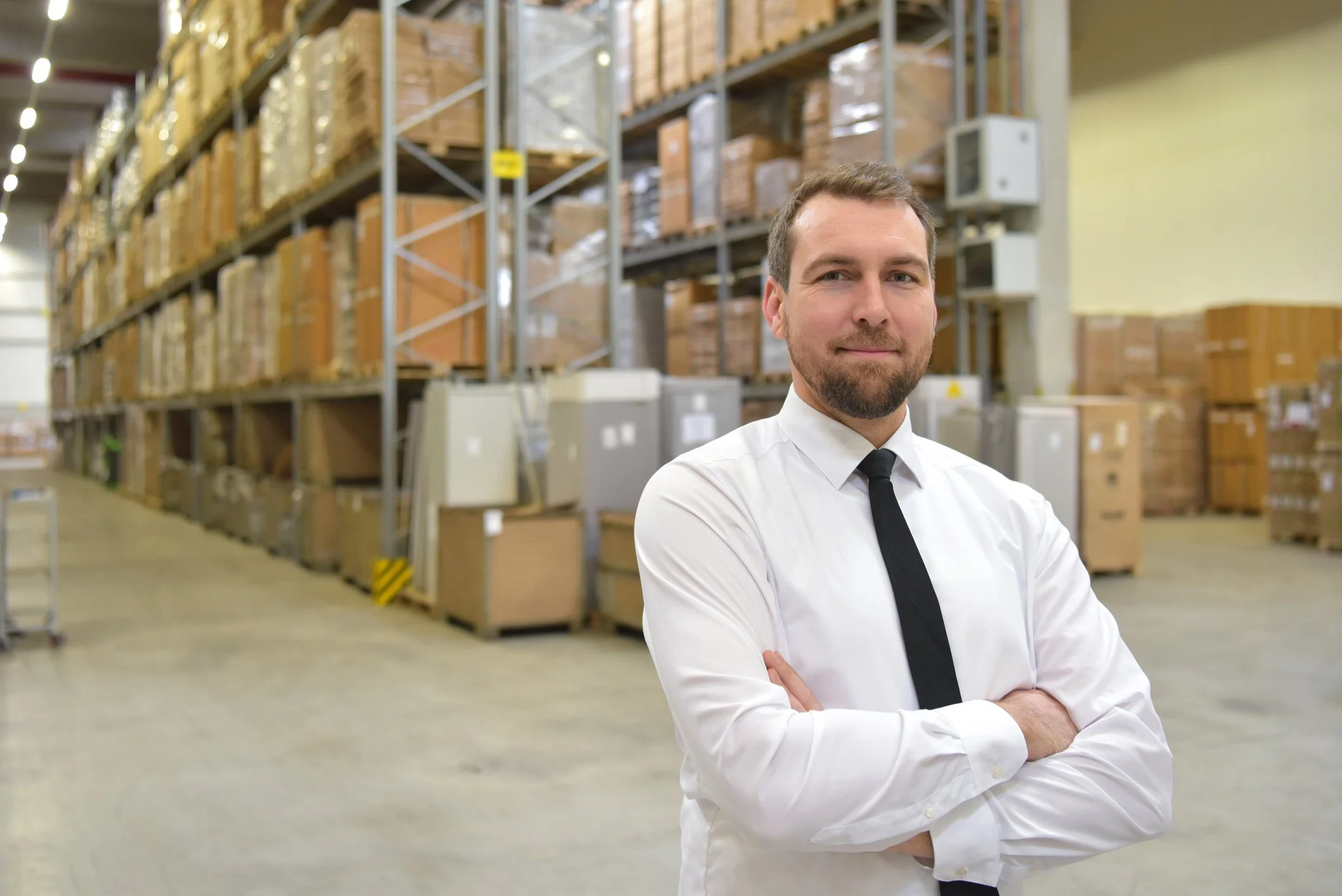 A smiling man in a white dress shirt and black tie stands with arms crossed in a warehouse with tall shelves filled with boxes and pallets.