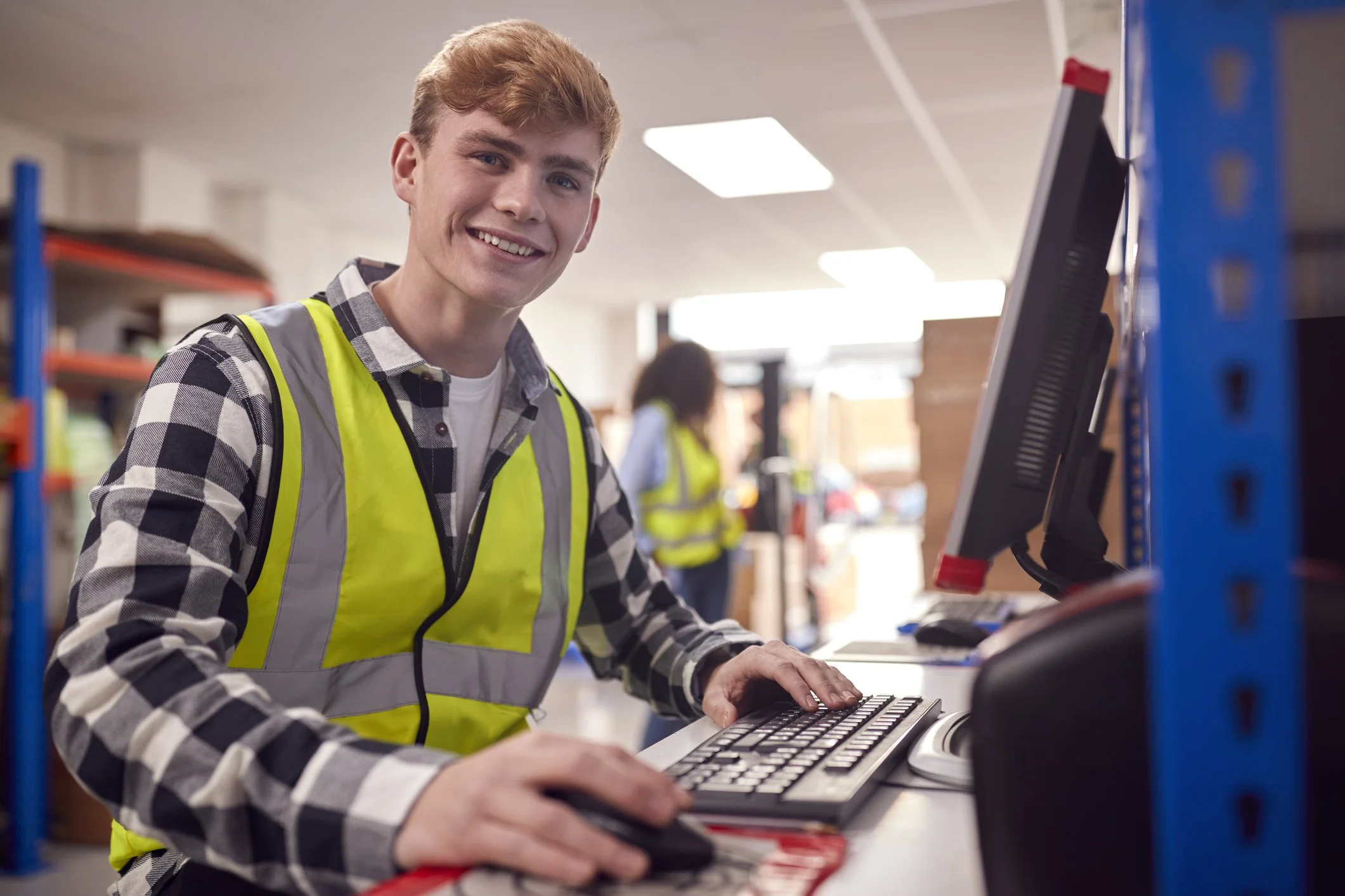 A young man in a checkered shirt and yellow safety vest working on a computer in a warehouse or office setting.