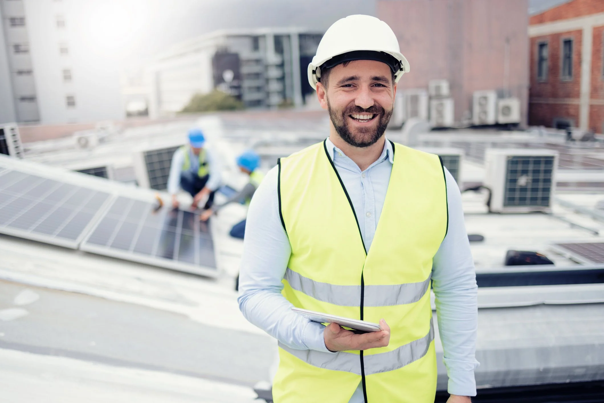 A smiling man wearing a white hard hat and a yellow safety vest, holding a clipboard, with solar panels and workers installing solar panels on a rooftop in the background.