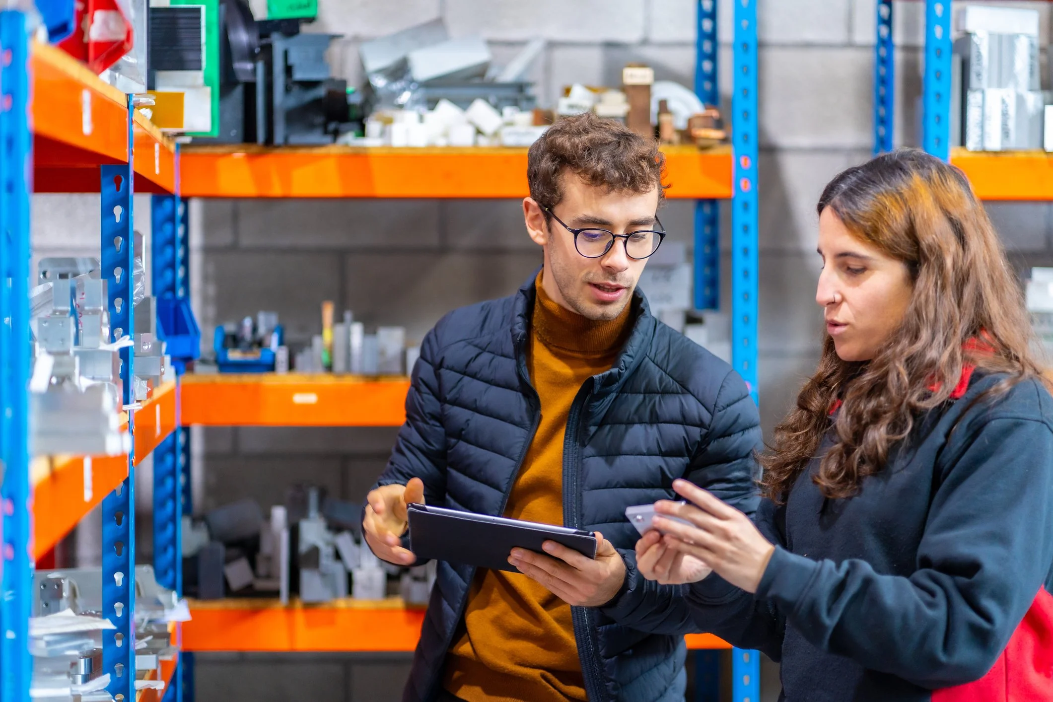 Two people are in a warehouse aisle, looking at a clipboard or tablet. The man has glasses, brown curly hair, and is wearing a navy puffer jacket and a mustard turtleneck. The woman has long, wavy brown hair and a nose piercing, and is wearing a dark jacket with a red shirt underneath. Shelves with boxes and hardware supplies are in the background.
