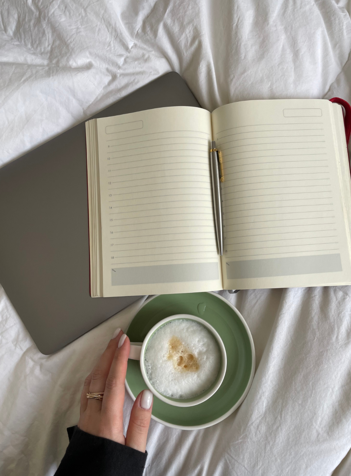 Open planner with lined pages and a silver pen resting on it, sitting on a closed gray laptop, with a person's hand holding a green saucer and cup of frothy cappuccino on a white bed.