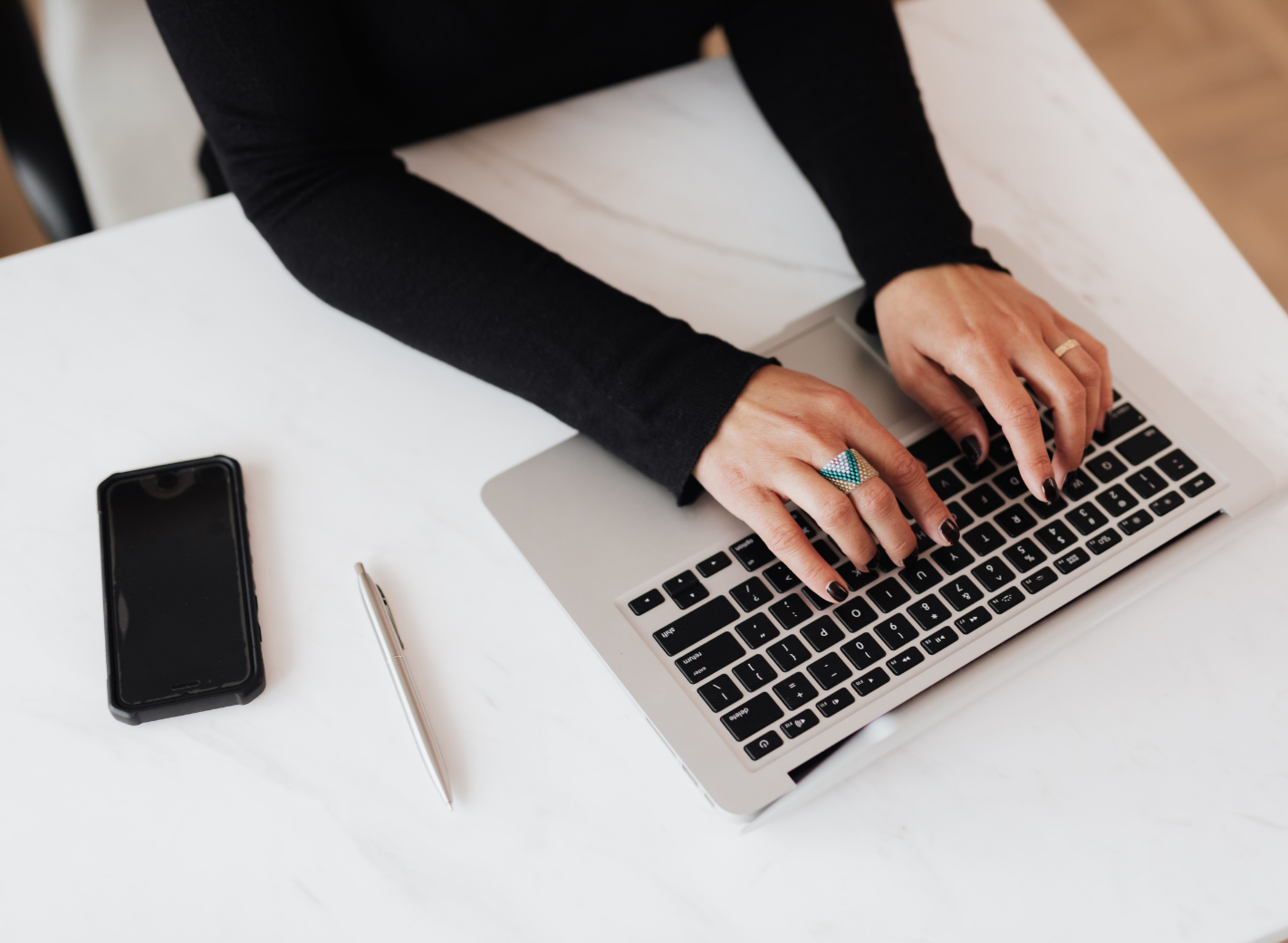 Person typing on a laptop with a smartphone, pen, and ring on their fingers on a white table.