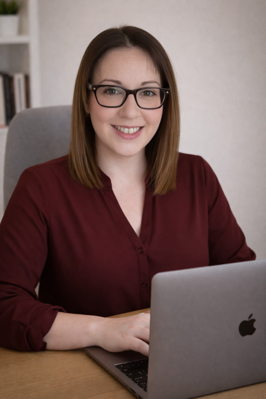 Picture of a woman wearing glasses at a desk smiling. Wearing a burgundy v-neck button blouse.