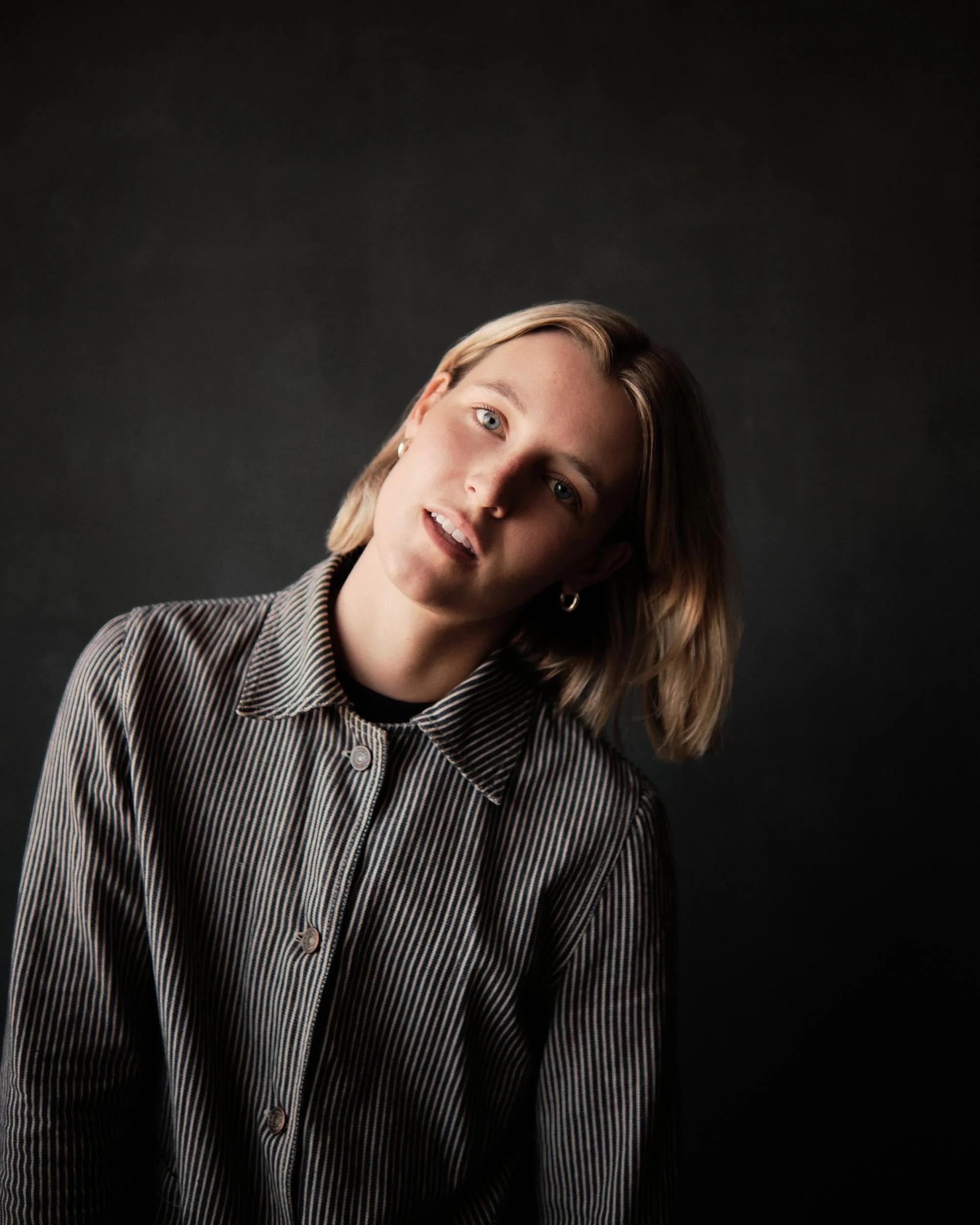 Portrait of a young woman with blonde hair and blue eyes, wearing a striped shirt, posing against a dark background.