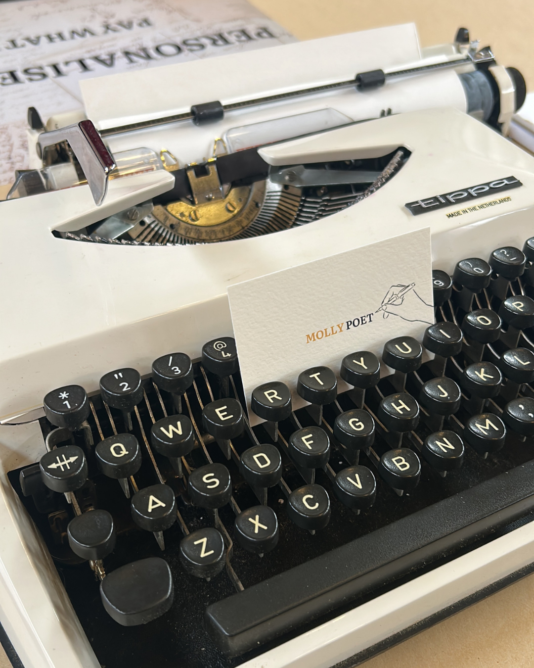 Close-up of a vintage white typewriter with black keys, a business card reading "MOLLY POET" placed on the keyboard, and printed paper in the paper tray, on a beige surface.