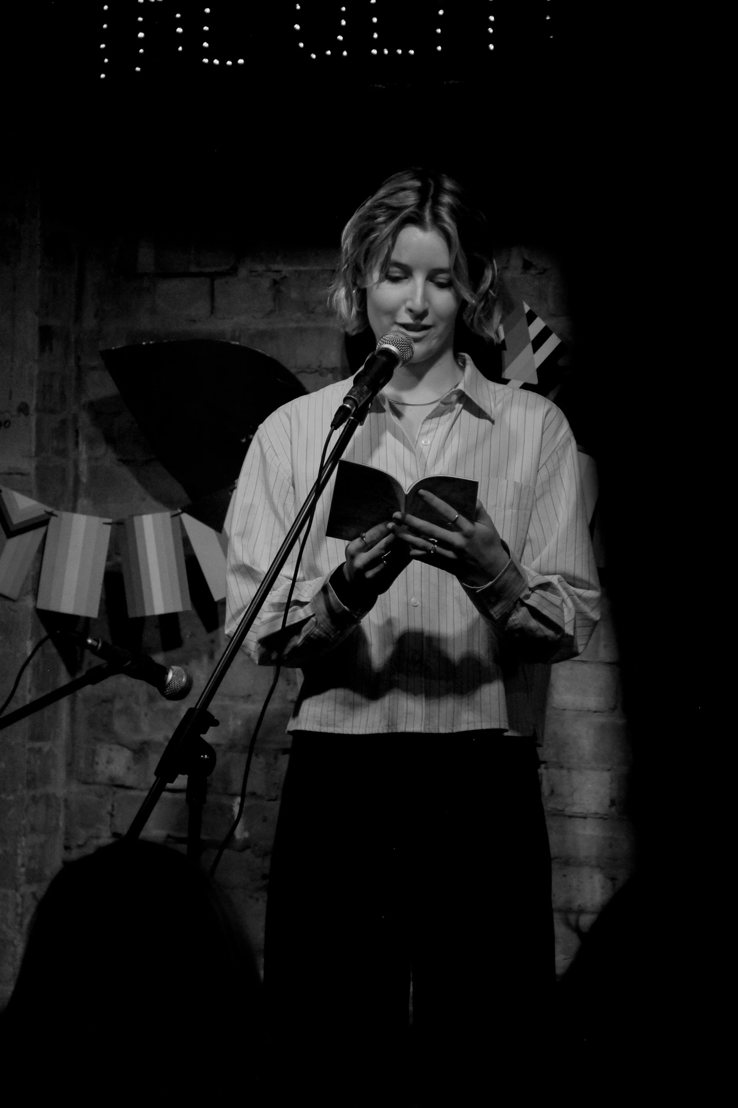 A young woman with short hair stands on stage reading from a book into a microphone, with a brick wall background and stage decorations.