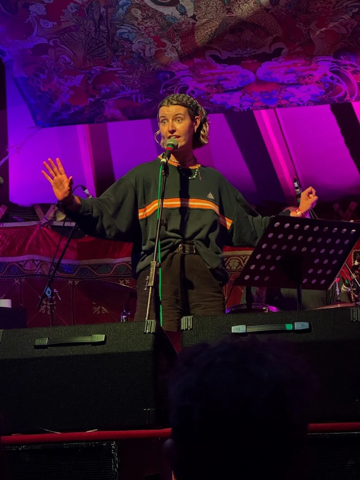 A woman performing on stage with coluorful lighting, wearing a dark Adidas sweatshirt with orange and yellow stripes, and gesturing with both hands.