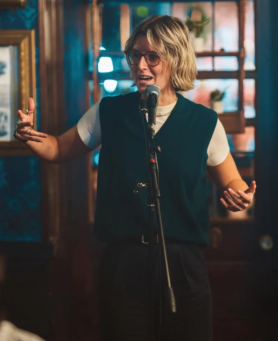 A woman with short blonde hair, glasses, and wearing a black vest over a white shirt, speaking into a microphone with one hand gesturing, in a cozy indoor setting with wooden shelves and potted plants.
