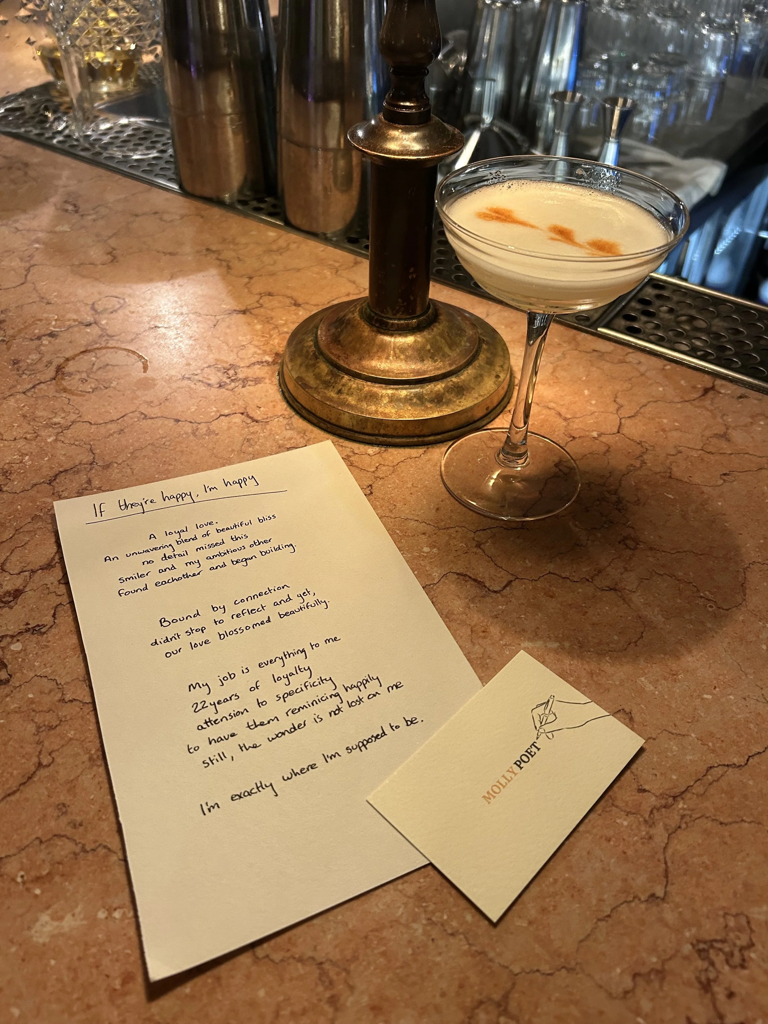 A cocktail glass containing a creamy drink with orange garnish, placed on a pinkish marble bar counter next to a handwritten poem and a small card with a drawing of a hand writing, labeled 'Molly Poem'. In the background, there are bar tools and bottles.