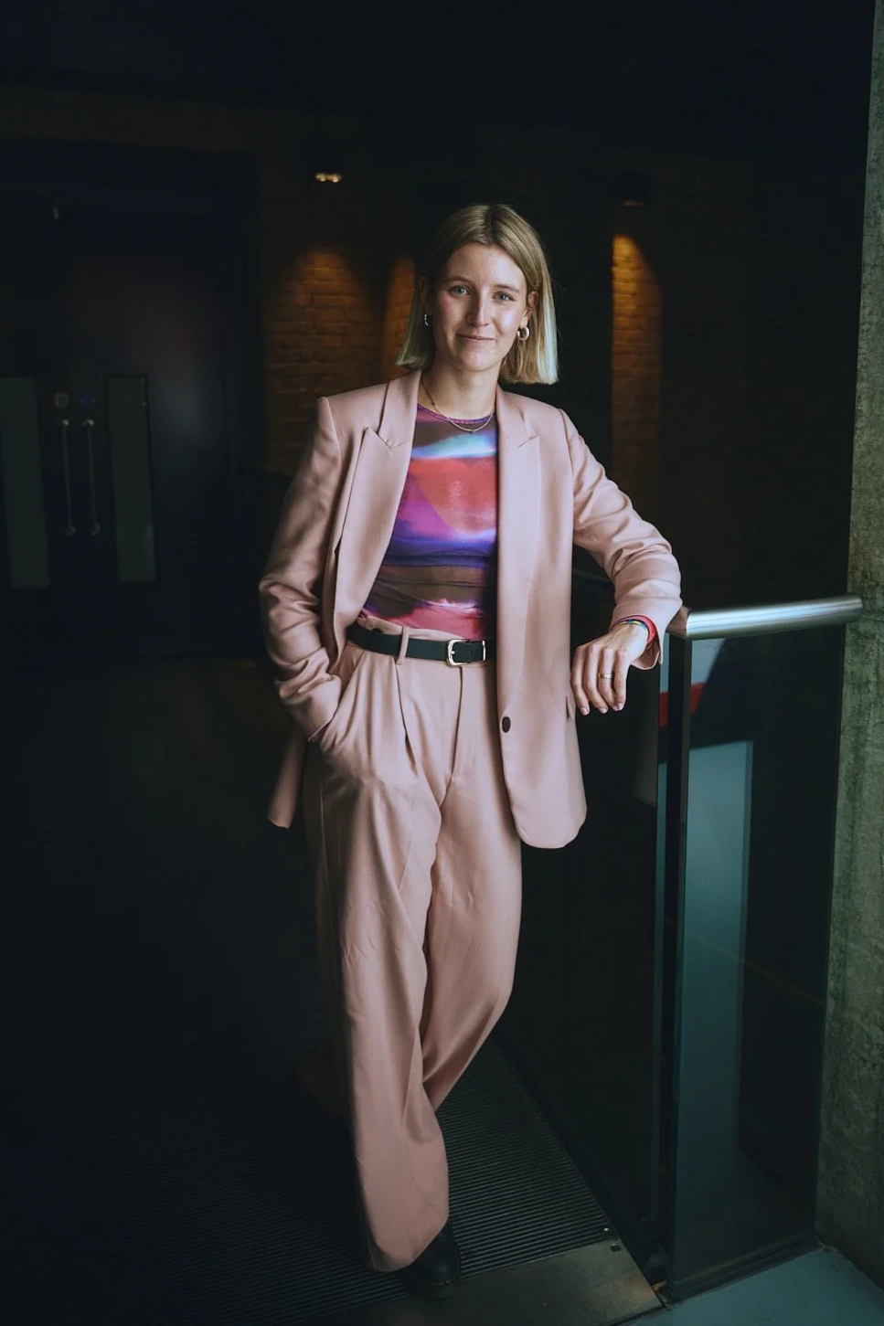 A woman with shoulder-length blonde hair wearing a pink suit and patterned top, standing indoors near a railing with a brick wall and dim lighting in the background.