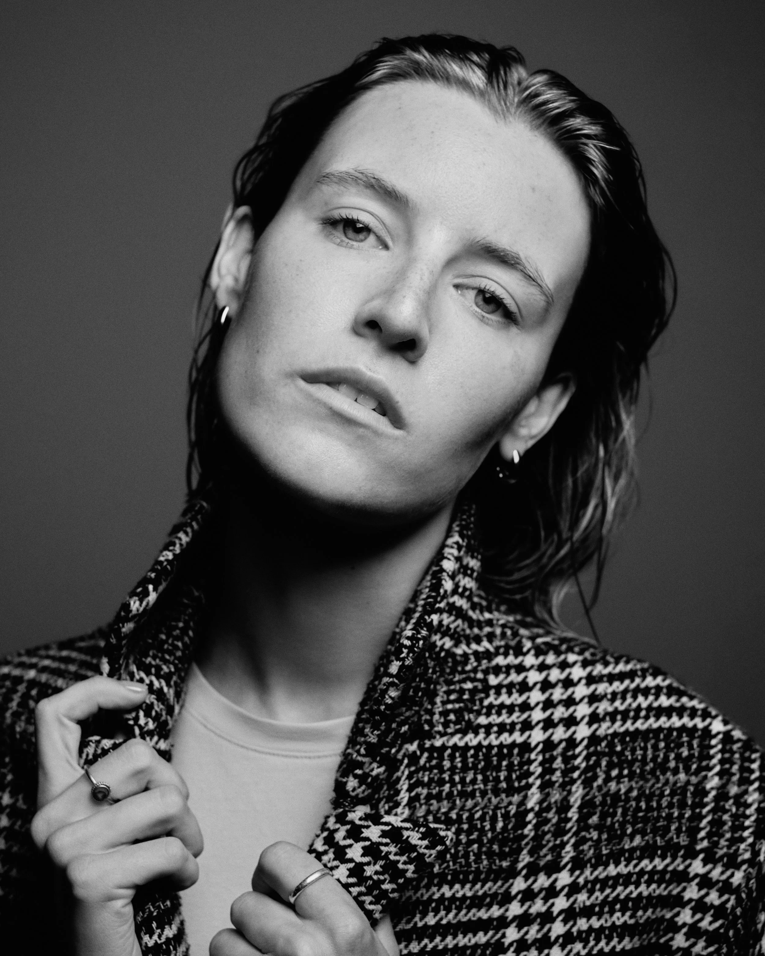 Black and white portrait of a woman with short hair, wearing a houndstooth patterned jacket and rings, looking at the camera.