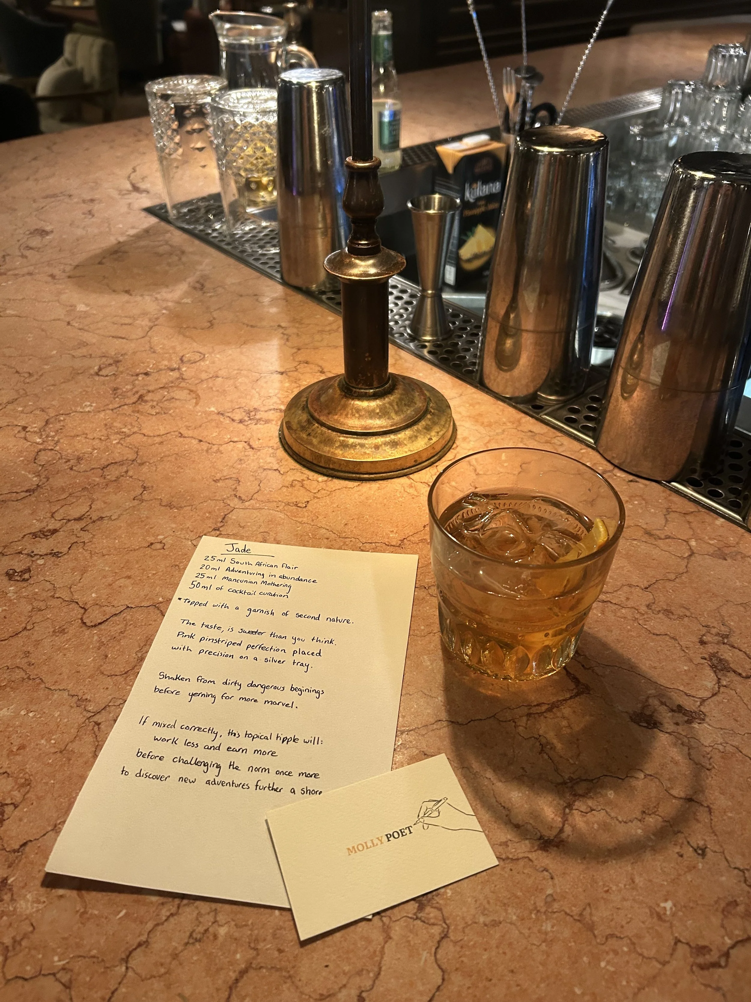 A bartender's counter with a brass candle holder, a glass of iced drink with lemon, a small note with a poem titled 'Jade', and a business card reading 'Molly Poet'. In the background, there are bar tools and glasses.