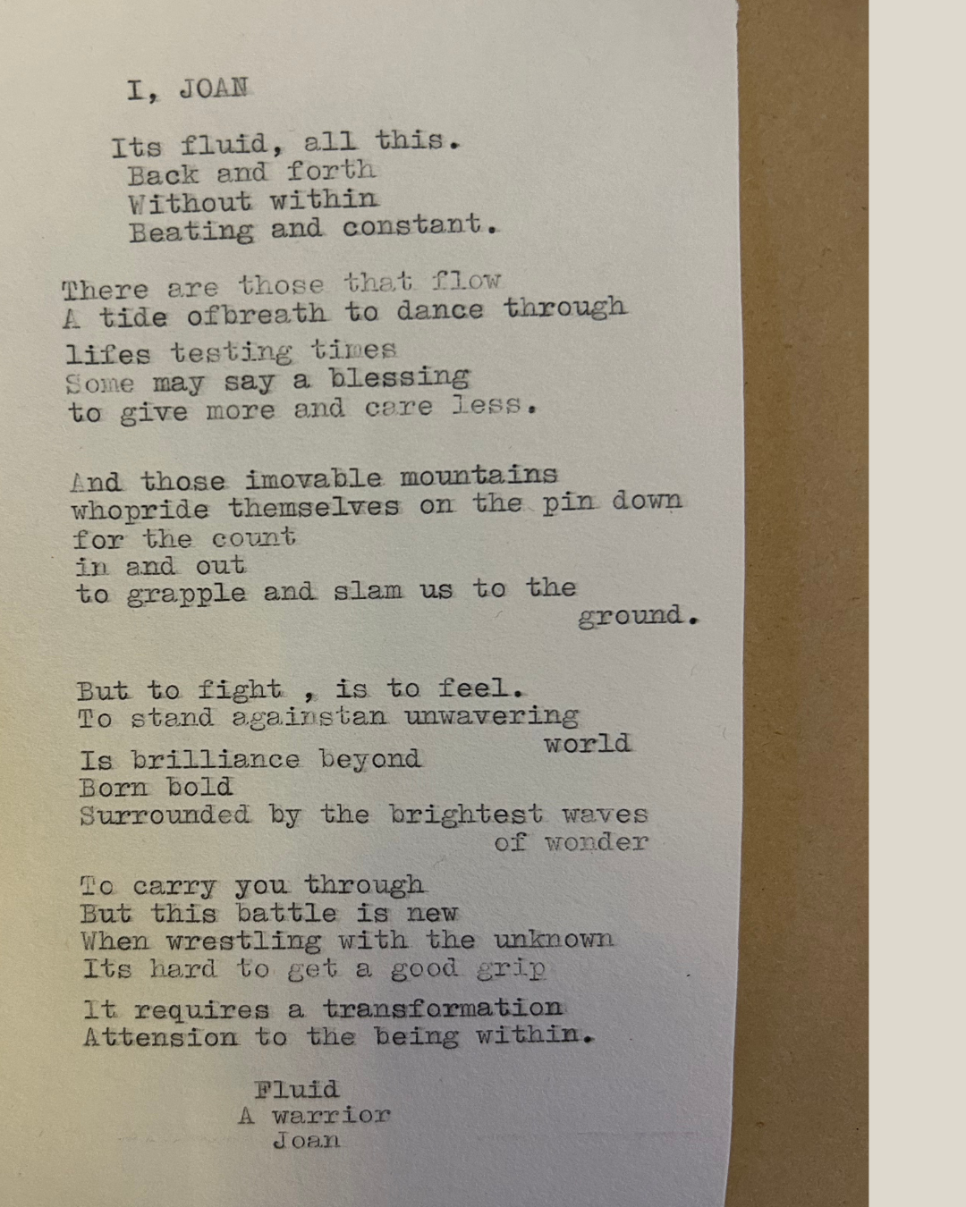 Typewritten poem on a white sheet of paper with a brown background. The poem is titled "I, JOAN" and discusses fluidity, mountains, and inner strength.