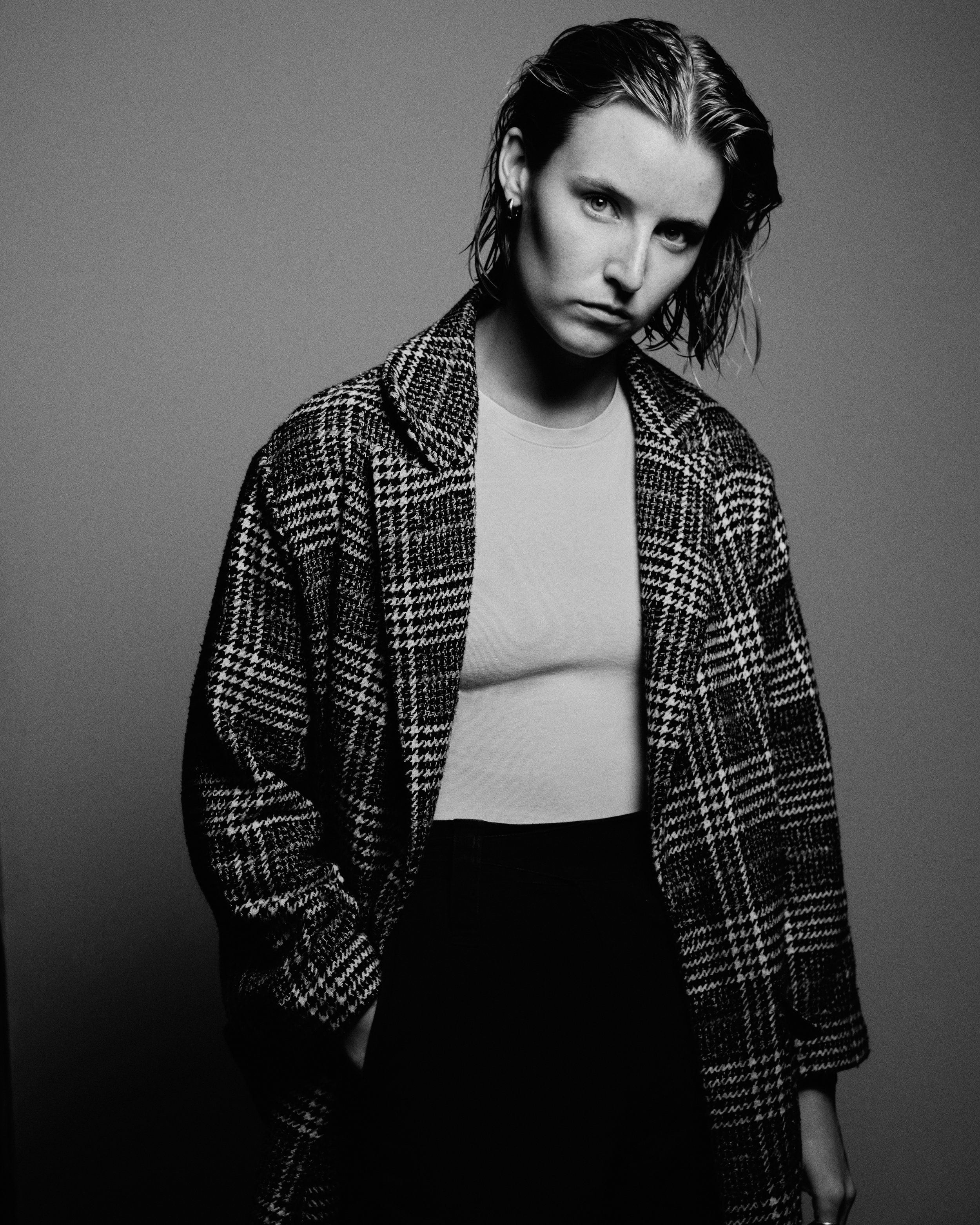 Black and white portrait of a woman with wet hair, wearing a patterned coat over a plain t-shirt, looking directly at the camera with a serious expression.
