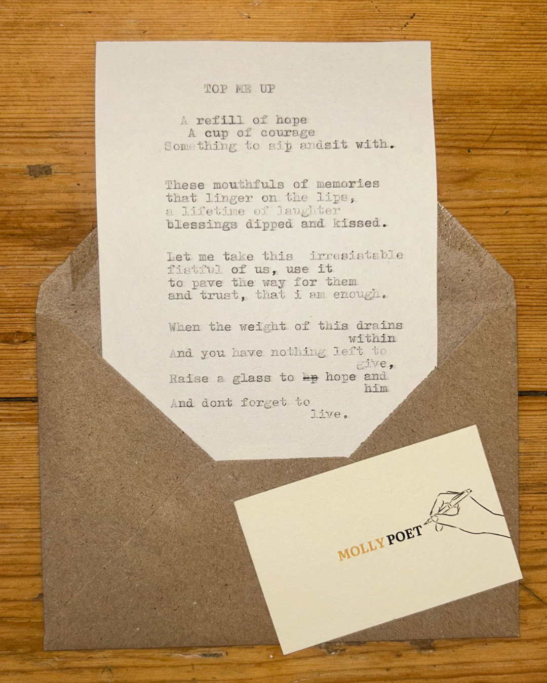 A handwritten poem titled 'Top Me Up' on a white paper, placed inside a brown envelope on a wooden surface, along with a small card that has 'MOLLY POET' printed on it with an illustration of a hand holding a pen.