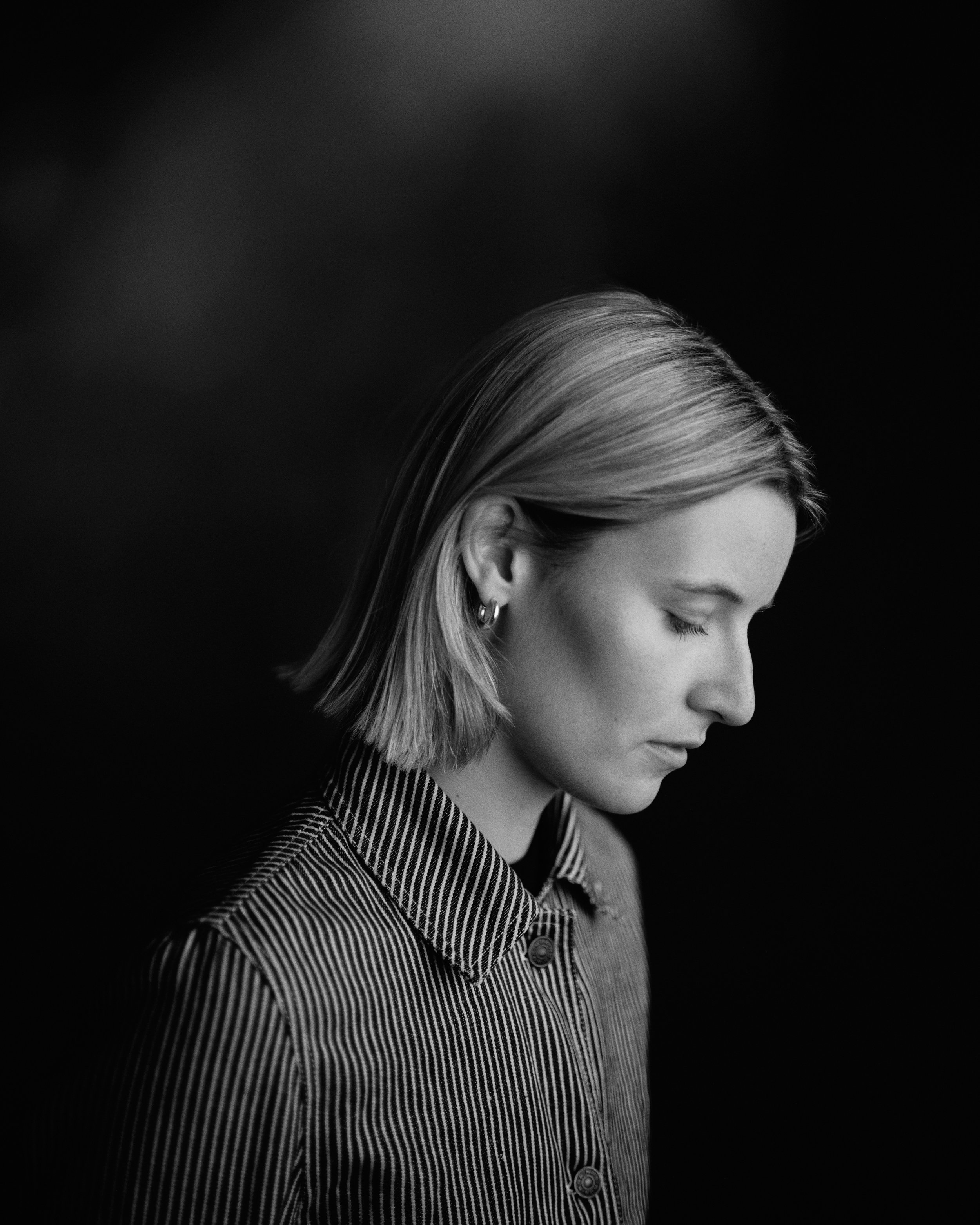 Black and white portrait of a young woman with short hair, wearing a striped collared shirt, with her eyes closed and her head tilted downward against a dark background.
