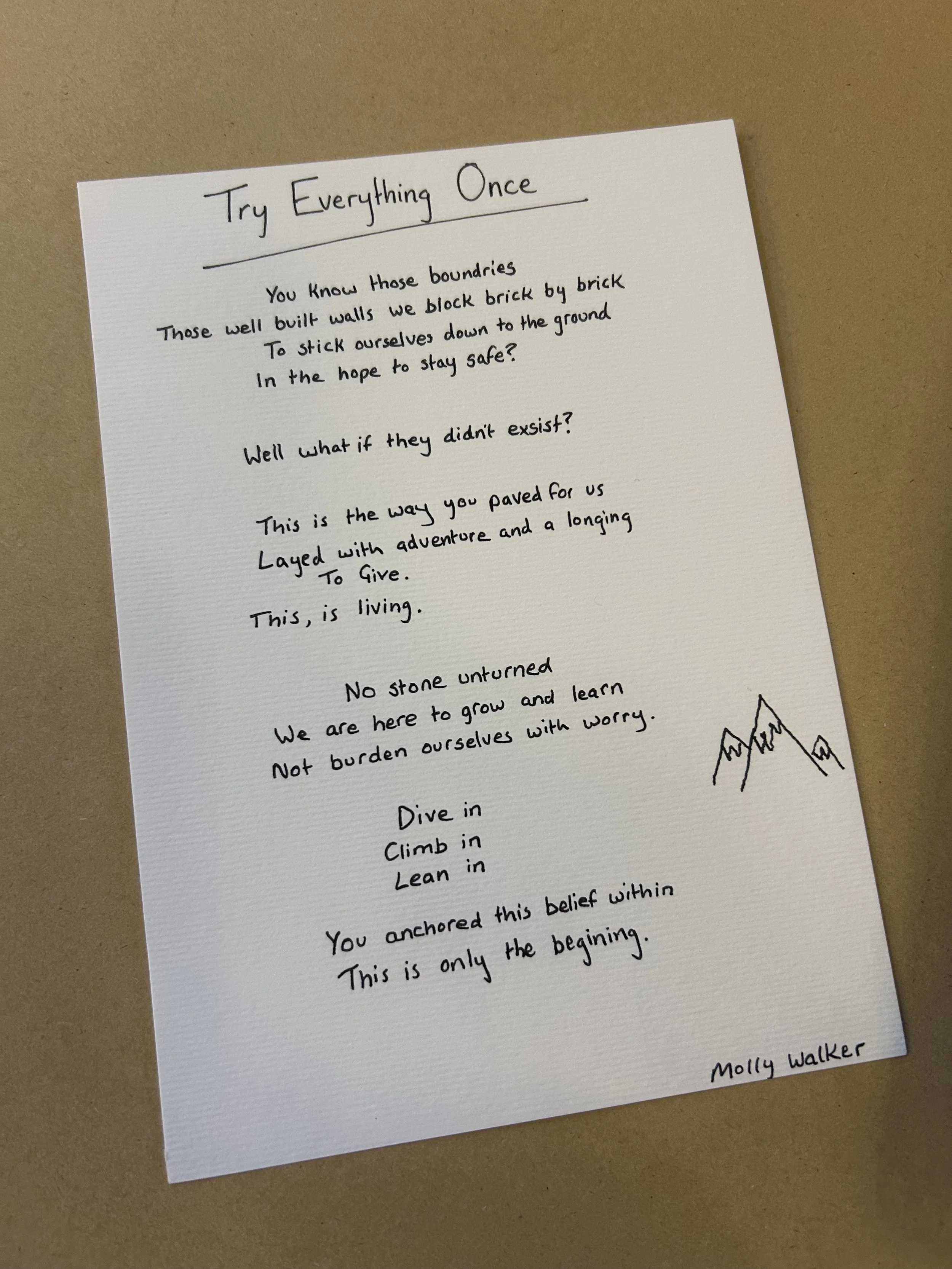 A handwritten poem titled 'Try Everything Once' on a white sheet of paper, placed on a brown surface. The poem is accompanied by a simple mountain sketch and a signature 'Molly Walker' at the bottom right corner.