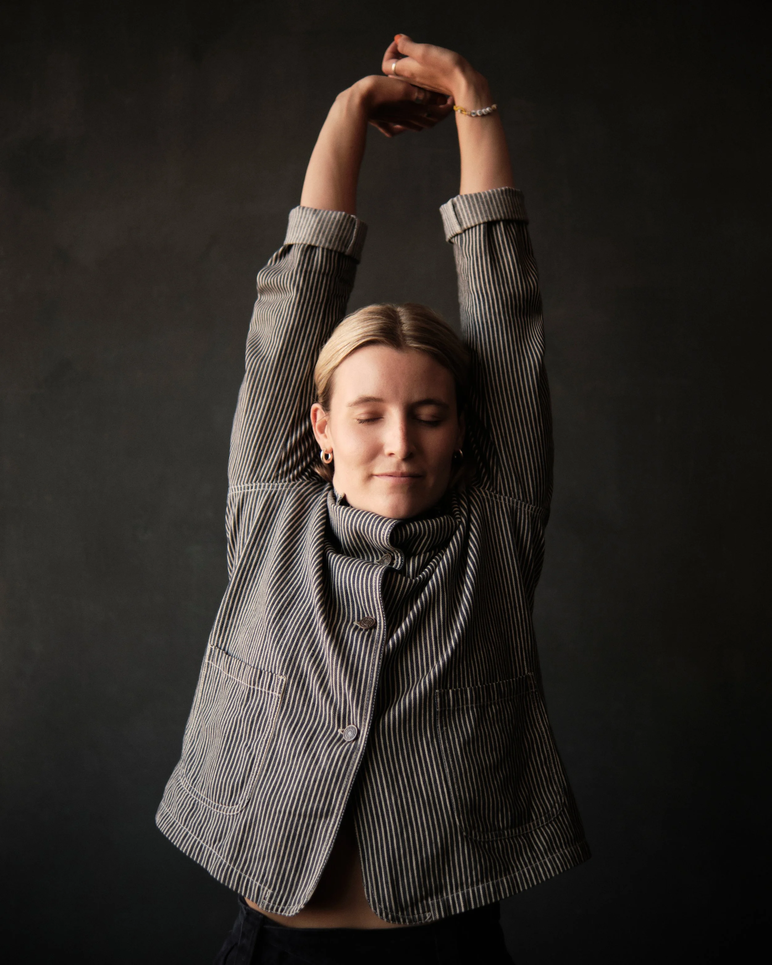 A woman stretching with her arms raised above her head, eyes closed, wearing a striped blazer against a dark background.