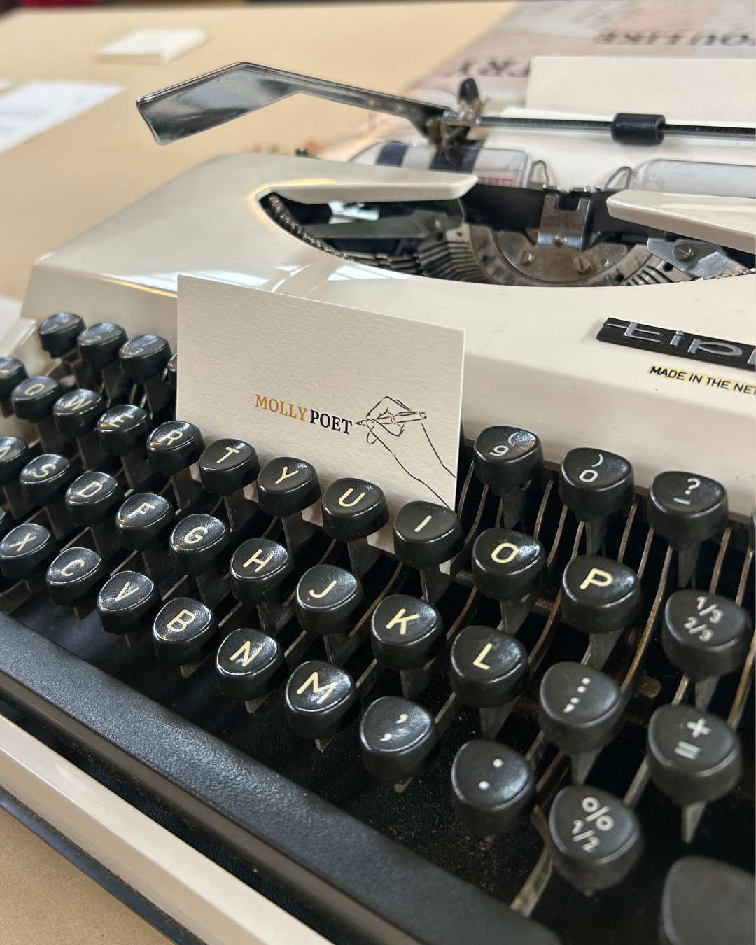 A vintage white typewriter with black keys, a blank note card inscribed with 'MOLLY POET' and a hand-drawn pen, and a driver’s license on a desk.