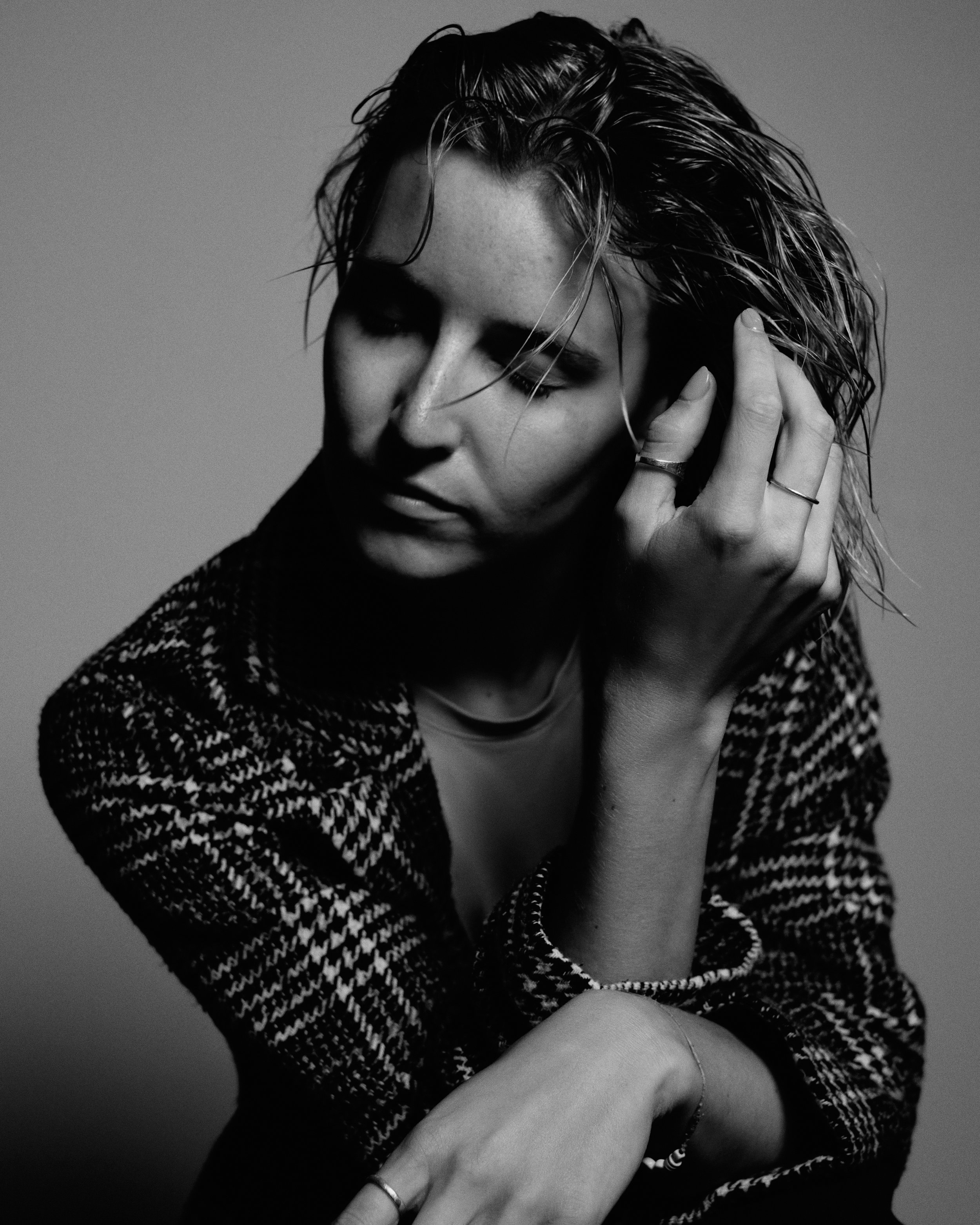 A black and white portrait of a woman with wet hair, closed eyes, and her hand touching her ear, wearing rings, a bracelet, and a patterned jacket.
