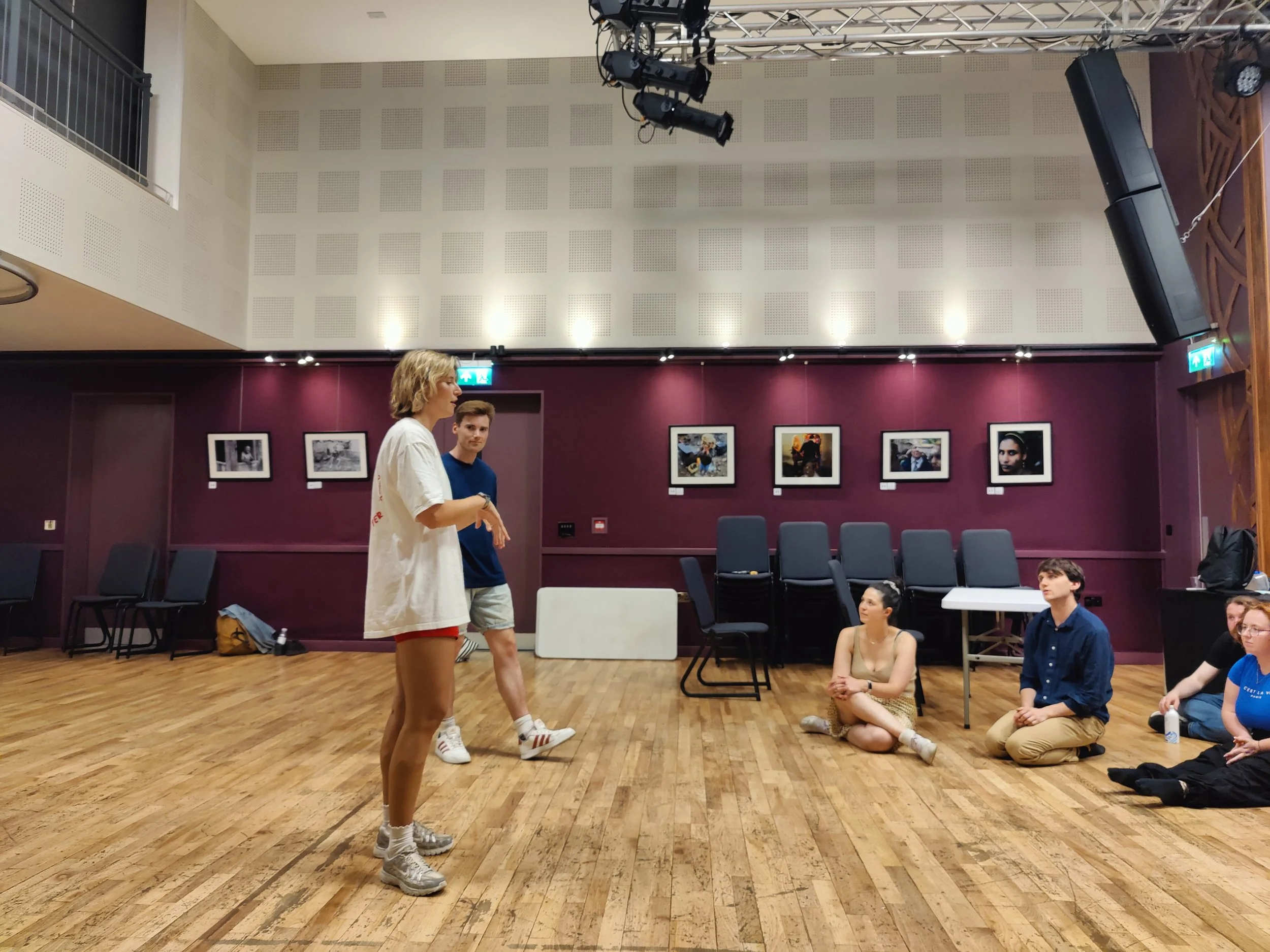 A group of people in a dance studio or rehearsal space with purple walls, hardwood floors, and phot0s hanging on the wall. Two individuals are standing, one speaking, while others sit on the floor listening.