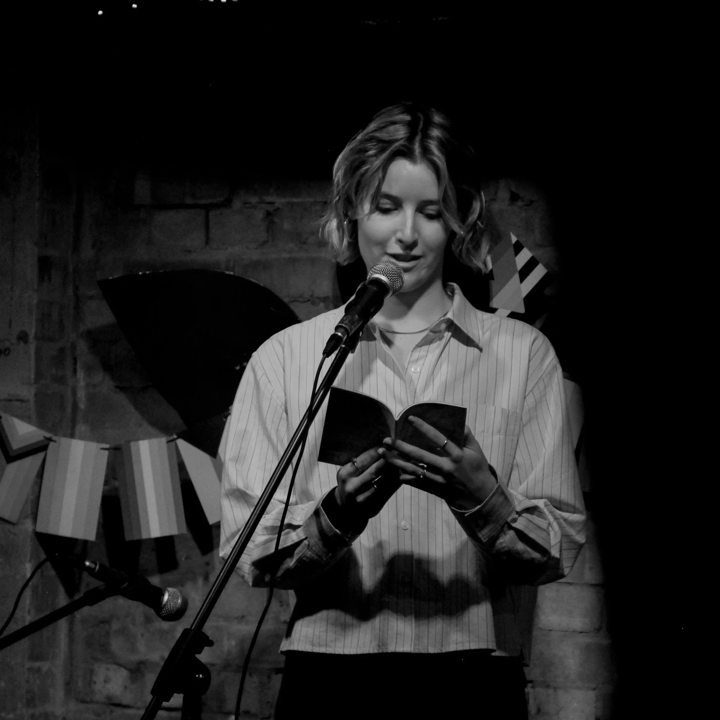 A woman with short wavy hair reading from a book into a microphone, standing against a brick wall with some decorations behind her.