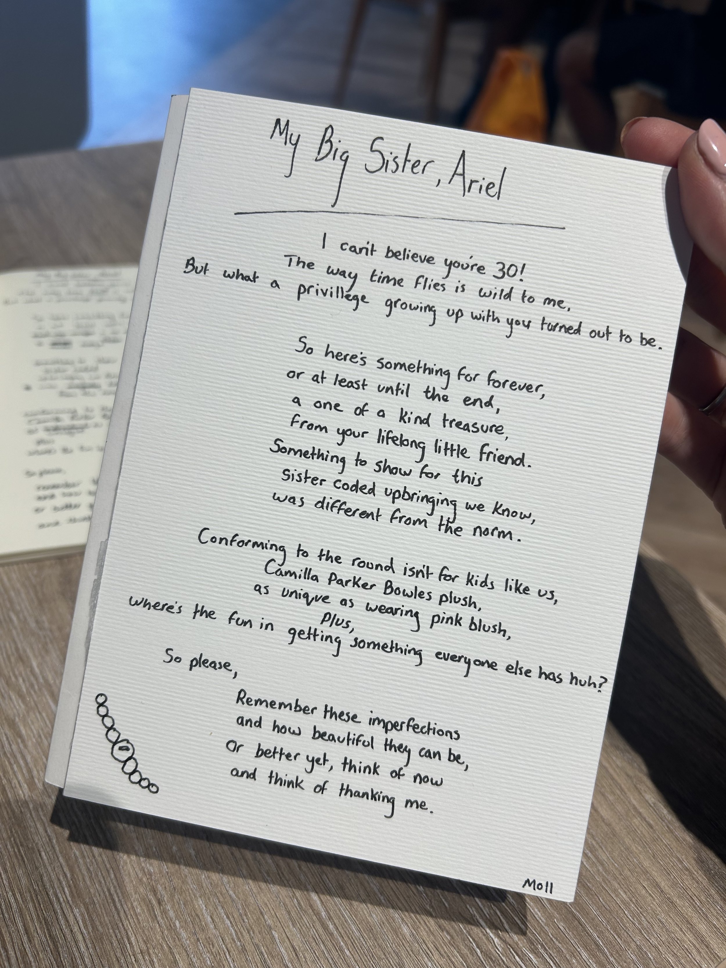 A handwritten note on white textured paper, held by a person with manicured nails, with text addressed to 'My Big Sister, Ariel'. The note expresses admiration and love, mentioning the importance of the sister, growing up together, and appreciation f