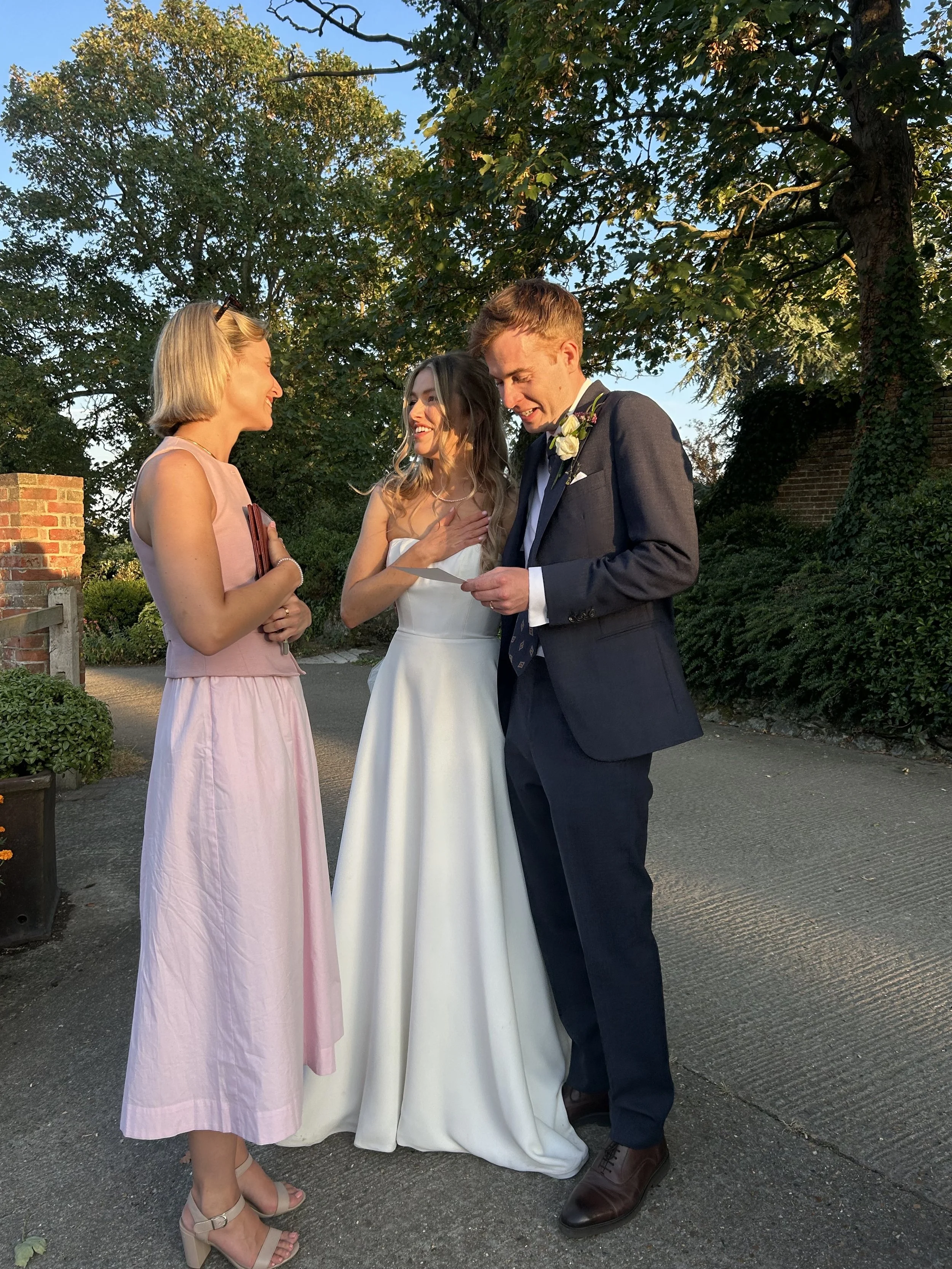 A bride and groom at a wedding ceremony outdoors, receiving a poem, written for them by Molly Walker in a pink dress. The scene is lit by sunlight, with trees and a brick wall in the background.