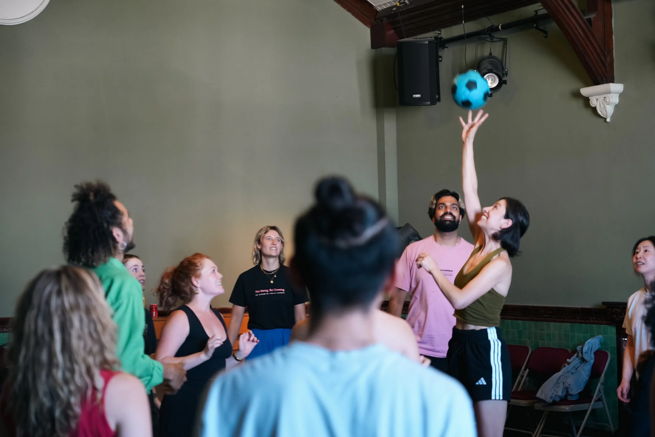 Group of people playing with a blue soccer ball indoors, some clapping and smiling, with a woman reaching up to hit the ball.