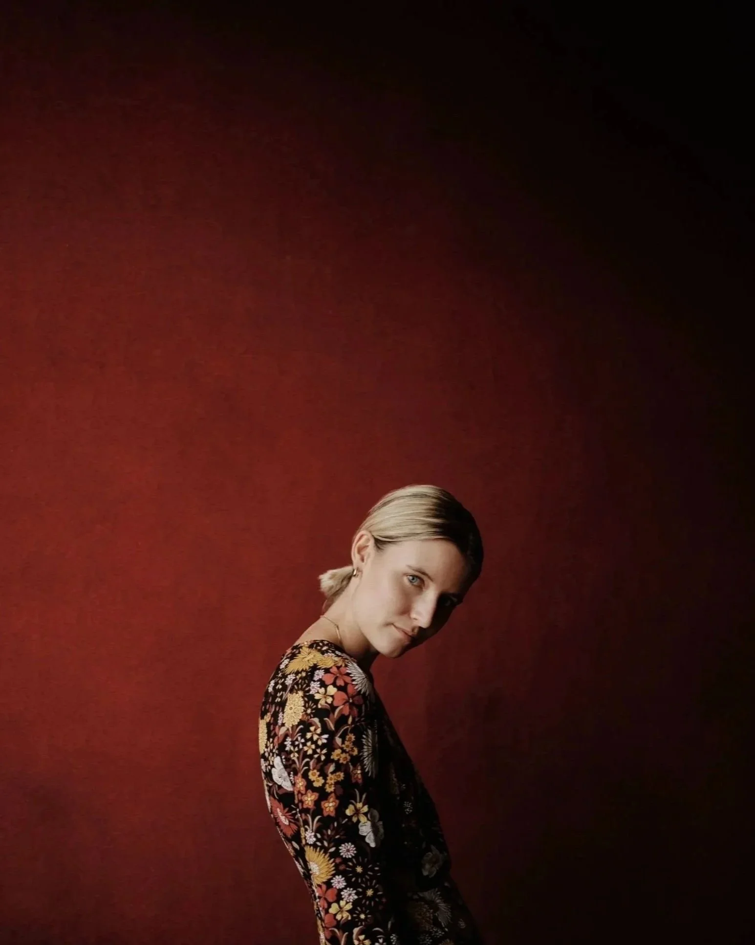 A woman with blonde hair tied back, wearing a floral print dress, standing against a red and black gradient background.
