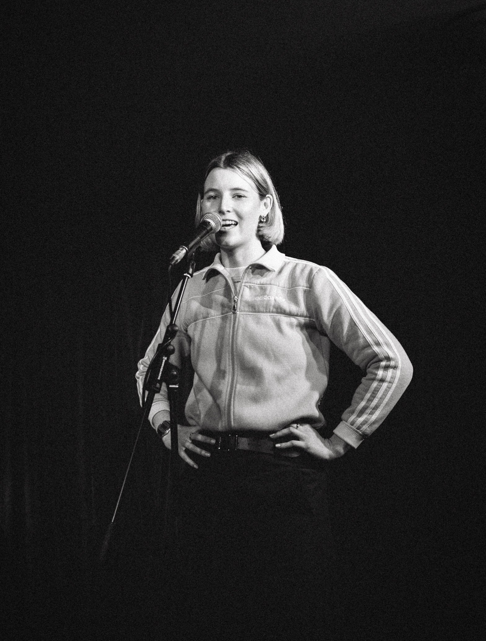 A young woman with short hair standing on stage, holding a microphone, and wearing a Adidas jacket.